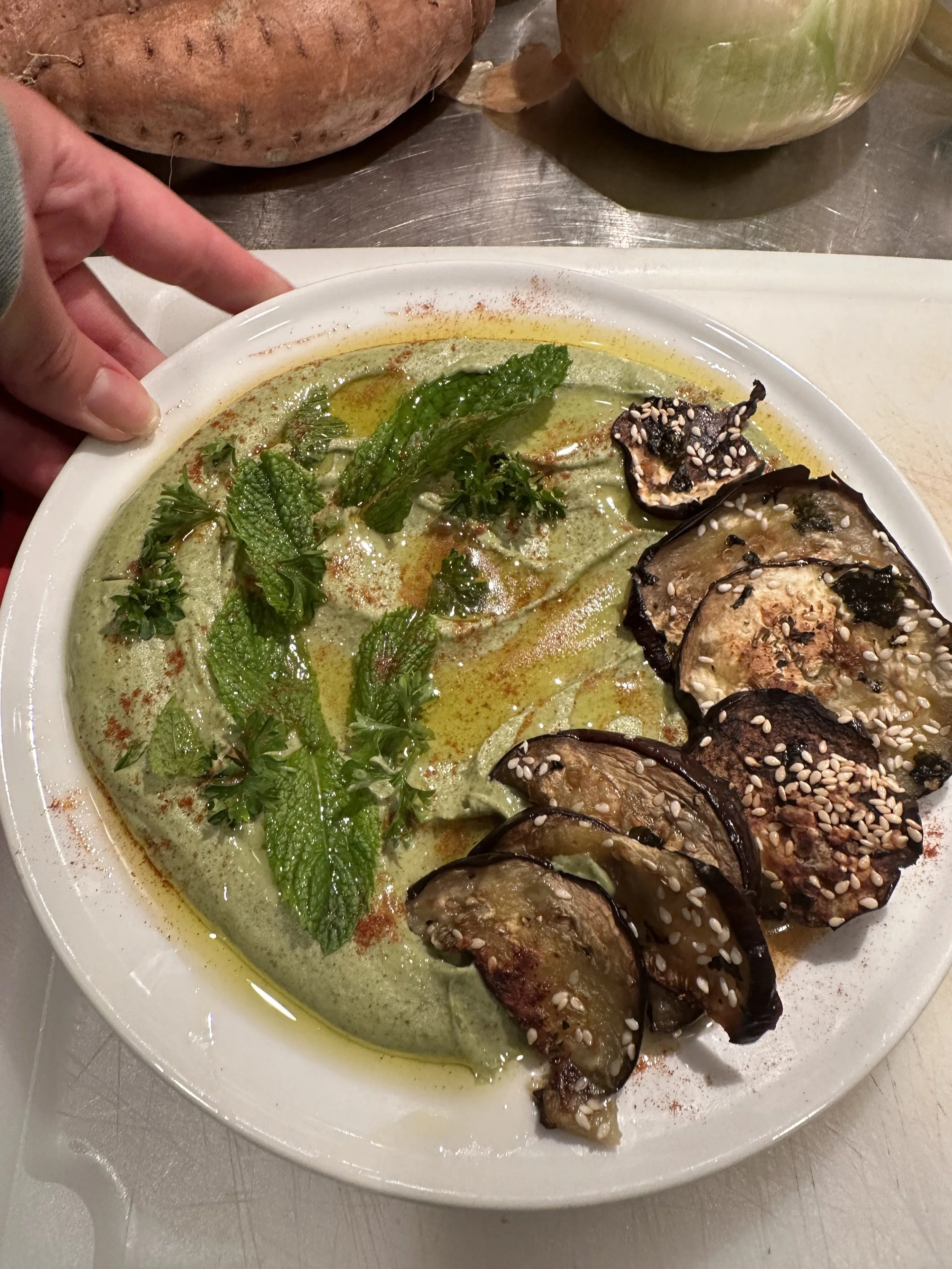 A plate of green-colored vegetable soup garnished with fresh herbs and oliated with a drizzle of oil, served with roasted eggplant slices topped with sesame seeds.