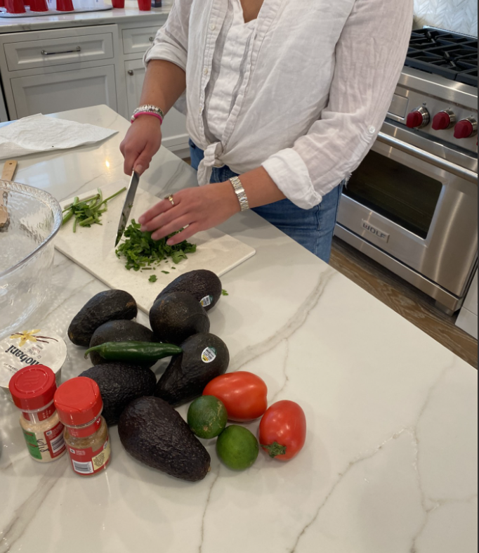 Person chopping cilantro on a white cutting board in a kitchen with avocados, tomatoes, limes, jalapeño, and spices on the counter.