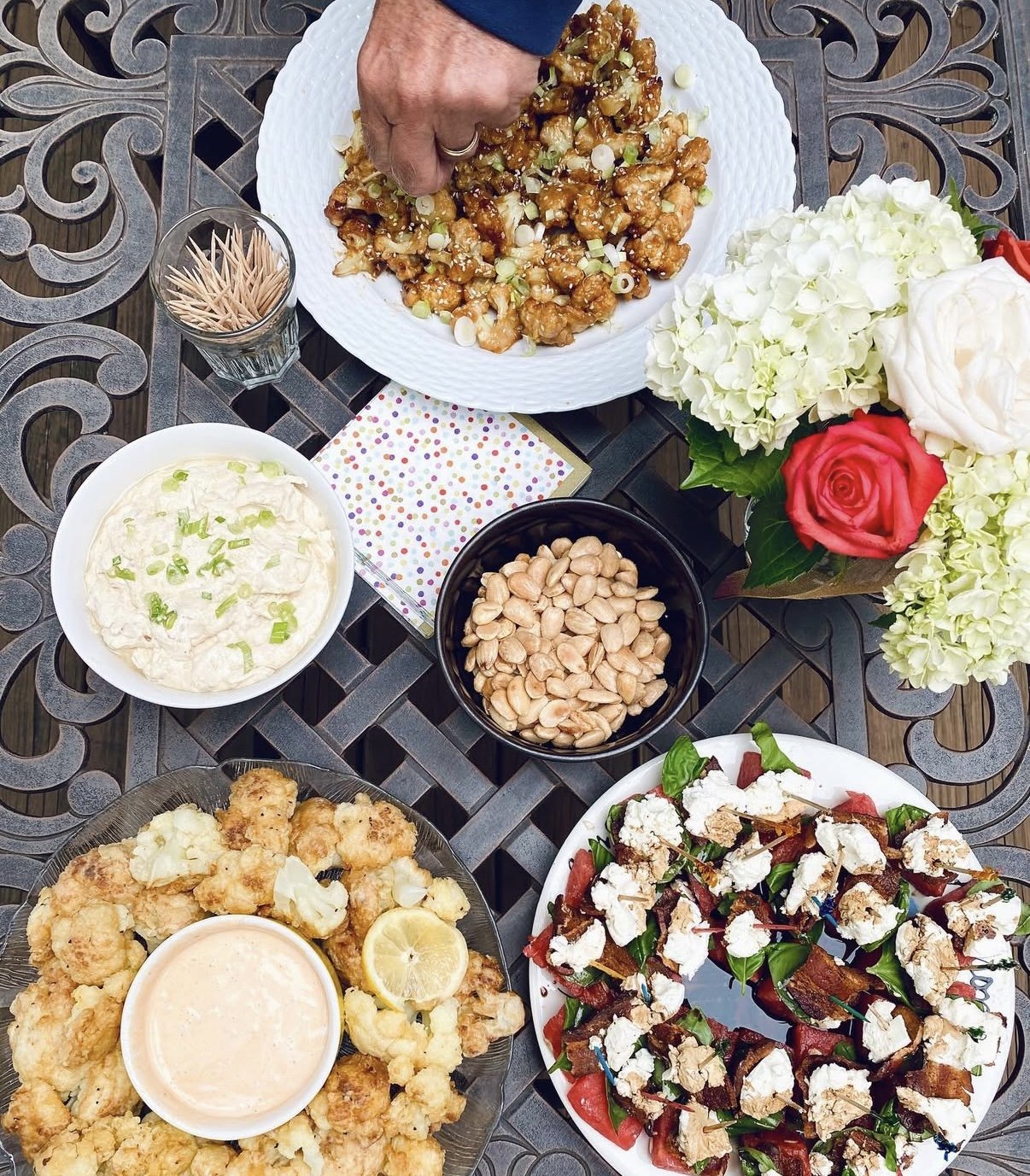 A table set for a meal with various dishes and flowers. There is a large plate of sweet and sour chicken, a bowl of potato salad, a bowl of peanuts, a platter of fried cauliflower with a dipping sauce, and a salad with tomatoes, spinach, and cheese. 
