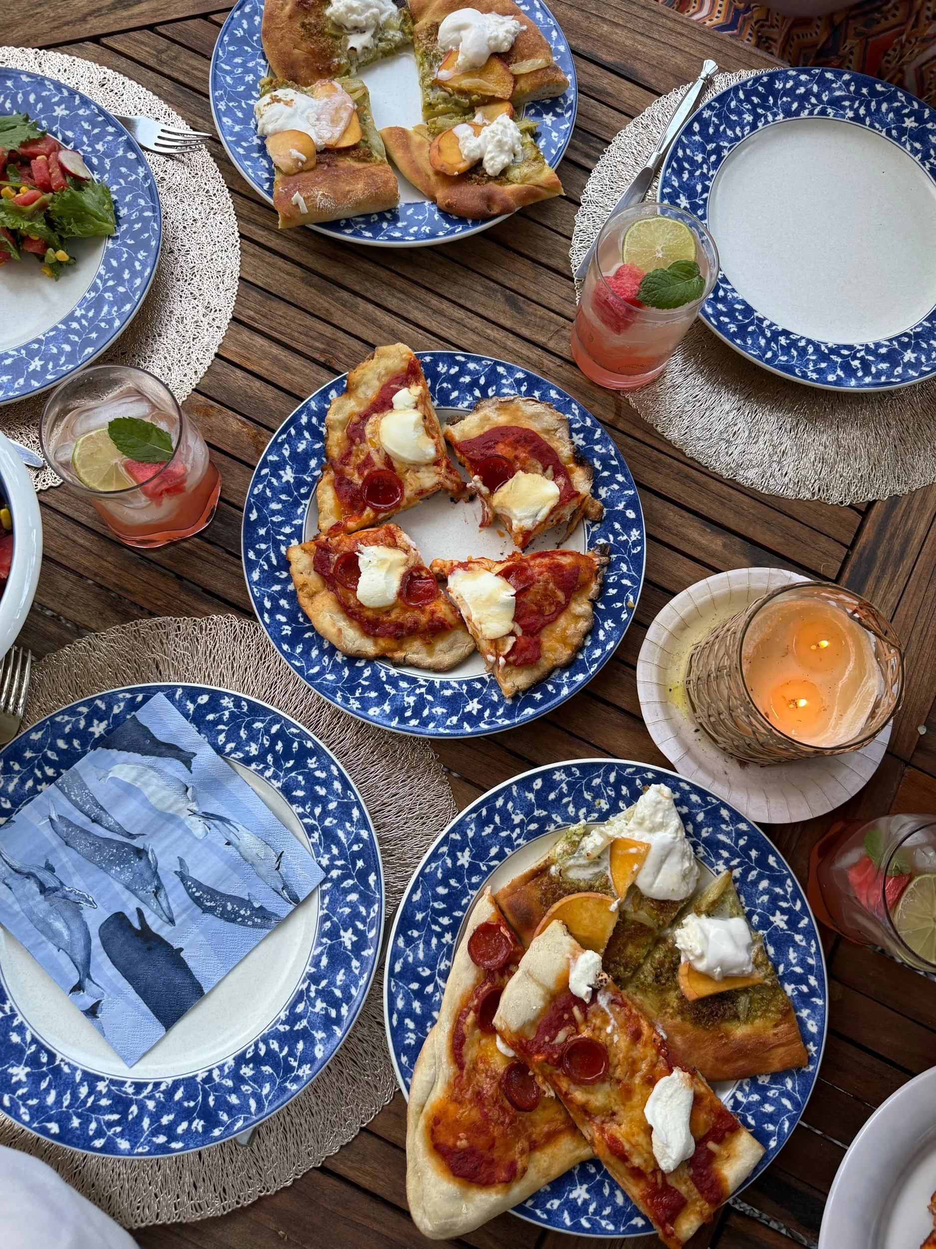 A table set with blue and white patterned plates filled with pizza slices, salad, and a variety of drinks, including water with lemon and mint, and pink beverages with fruit garnishes, on a wooden outdoor table with woven placemats and a candle