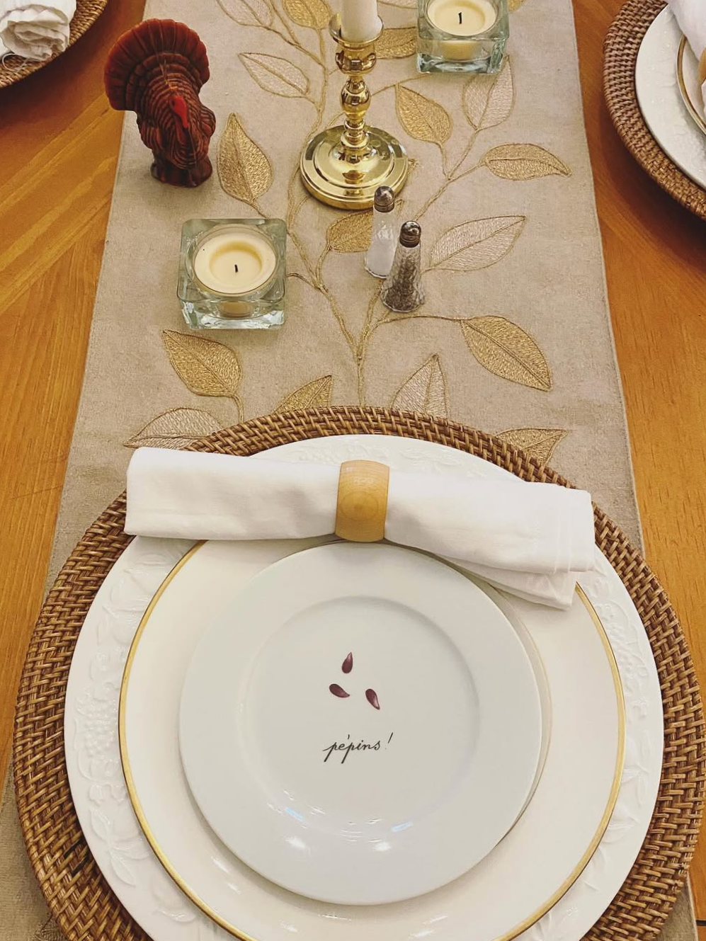 Elegant table setting with a white plate and a smaller bowl on top, embroidered with the word 'pépins!' and painted seeds, a rolled white napkin with a wooden napkin ring, decorative candles, salt and pepper shakers, and a table runner with gold embr
