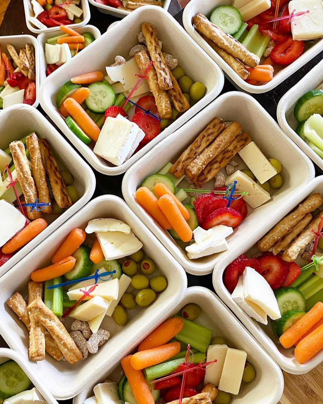 Multiple containers of colorful vegetable and cheese snacks with pretzel sticks, cherry tomatoes, cucumbers, carrots, and cheese slices.