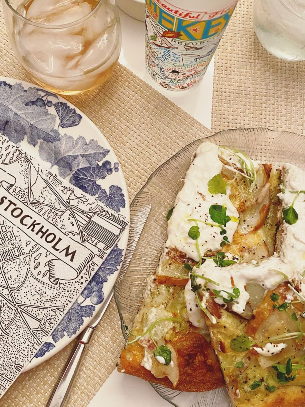 A table setting with a plate featuring a map of Stockholm, a glass of iced drink, a colorful travel-themed paper cup, and a glass dish with a slice of pizza topped with sauce, greens, and cheese.