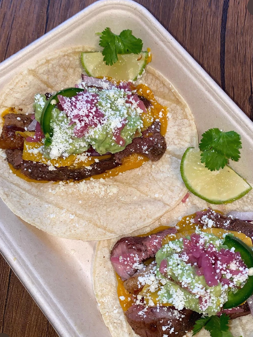 Two beef tacos on corn tortillas topped with shredded cheese, guacamole, sliced cucumbers, cilantro, lime wedges, and chopped onions, on a white platter.