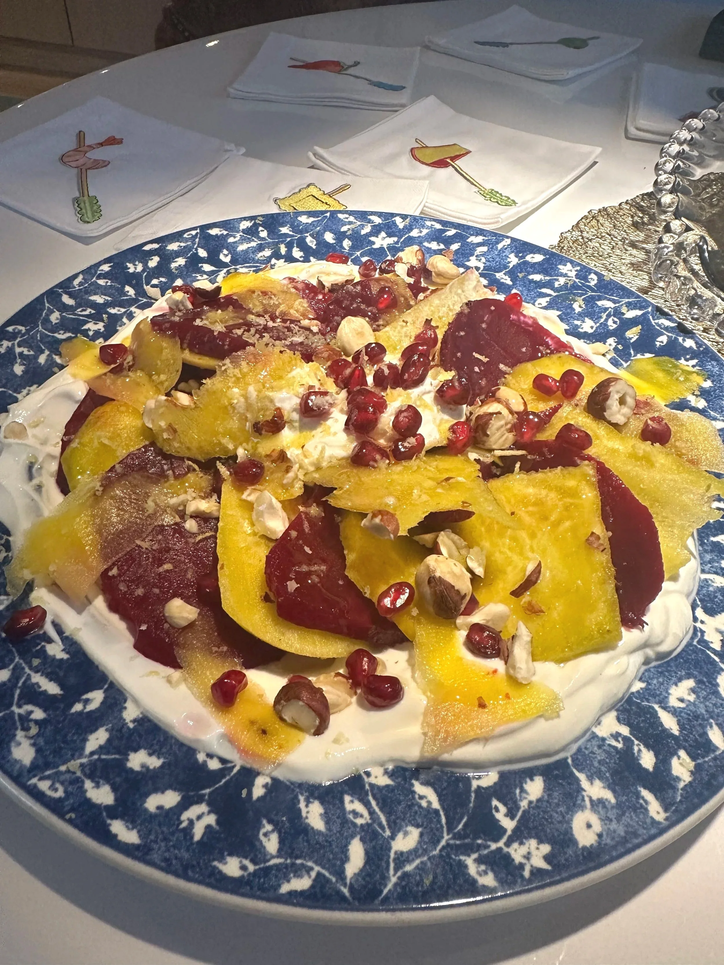 A large plate with a dessert that looks like a yellow and red fruit salad with pomegranate seeds and chopped nuts on top, served on a blue and white decorative plate.