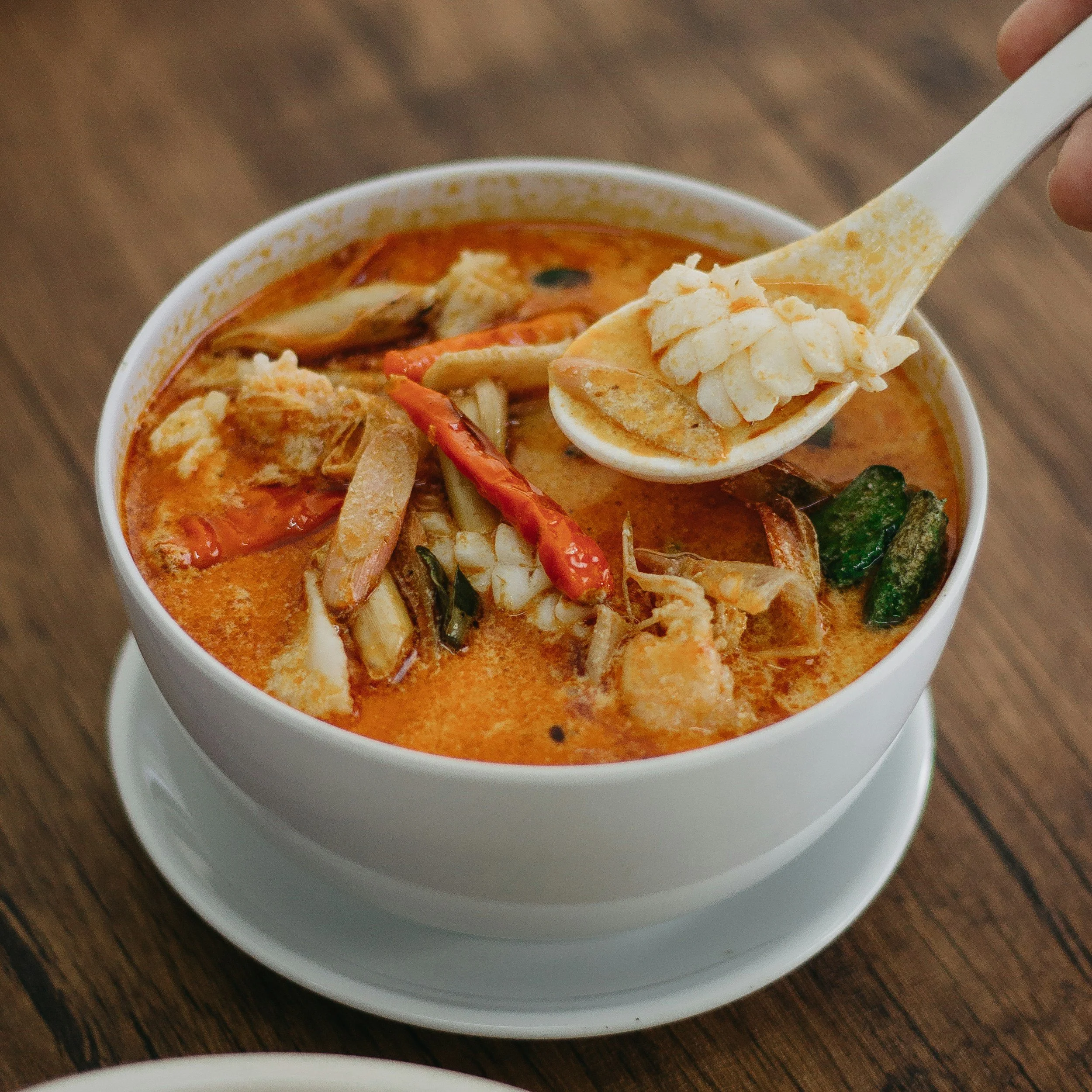 A bowl of spicy Thai red curry with vegetables and seafood, with a spoon holding rice, on a wooden table.