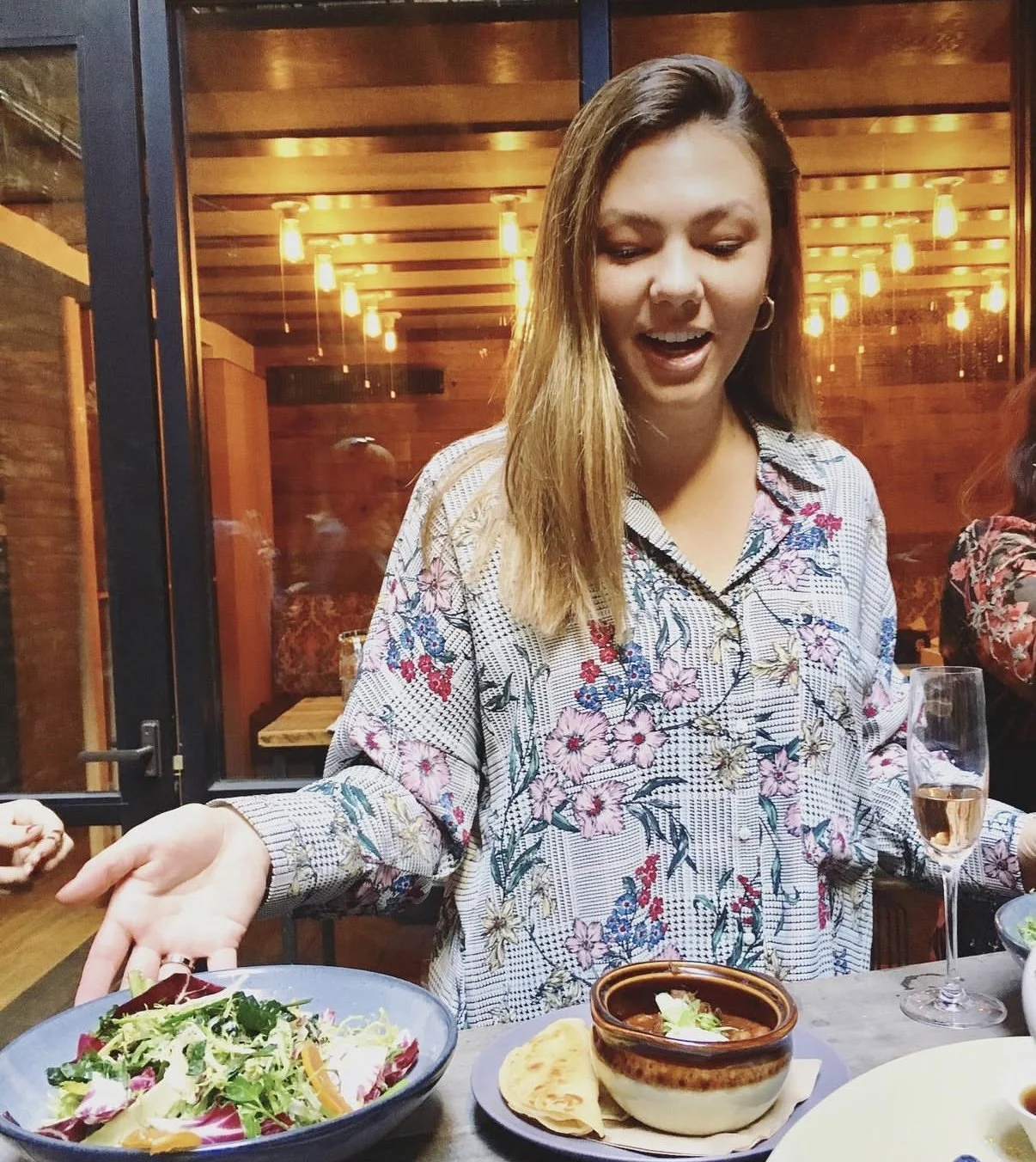 A woman in a floral shirt is serving herself salad from a bowl and has a bowl of soup with a piece of bread and a glass of rosé wine on the table in front of her.