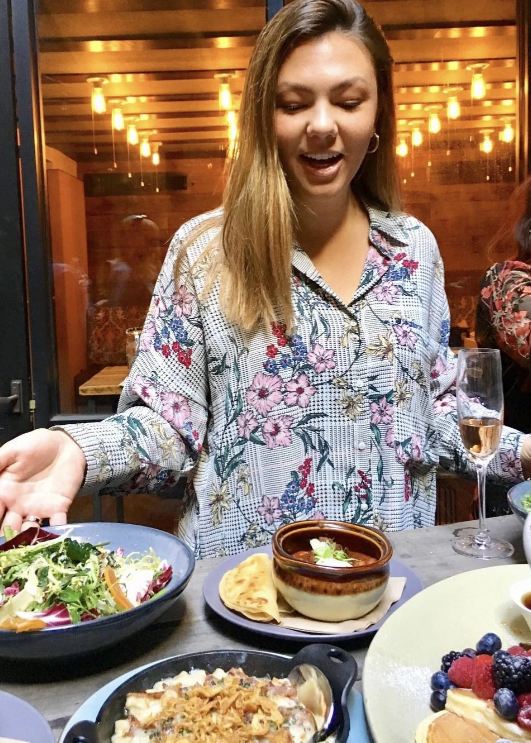 A woman in a floral patterned shirt standing behind a table of food, including a bowl of salad, a bowl of soup with bread, a skillet of baked pasta, and a plate of berries with pancakes, in a warmly lit restaurant.