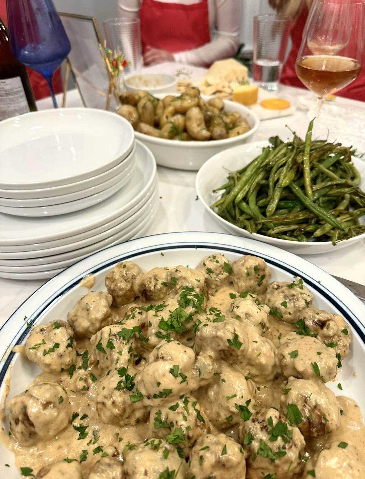 A family dinner table with a white plate of meatballs in creamy sauce topped with parsley, green beans, roasted root vegetables, wine glass with rosé, water glass, and stacked empty bowls.