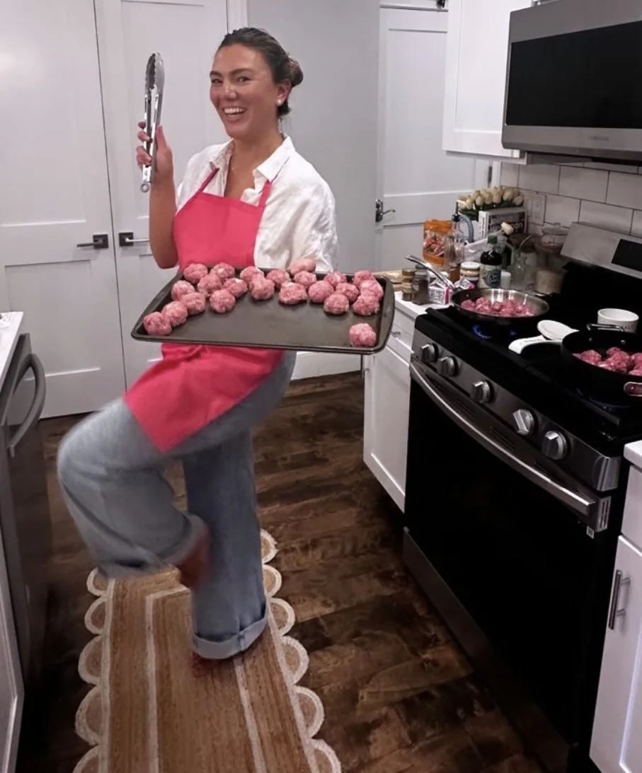 A woman in a white shirt and pink apron in a kitchen, holding a baking sheet with raw meatballs, smiling at the camera.