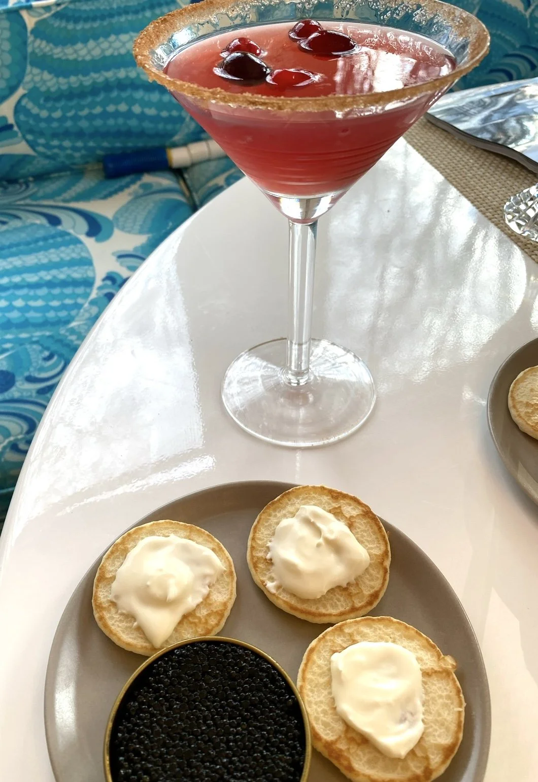 A martini glass filled with a red cocktail garnished with cherries and a salted rim, placed on a white table next to a plate with cream cheese topped crackers and a small dish of black caviar.