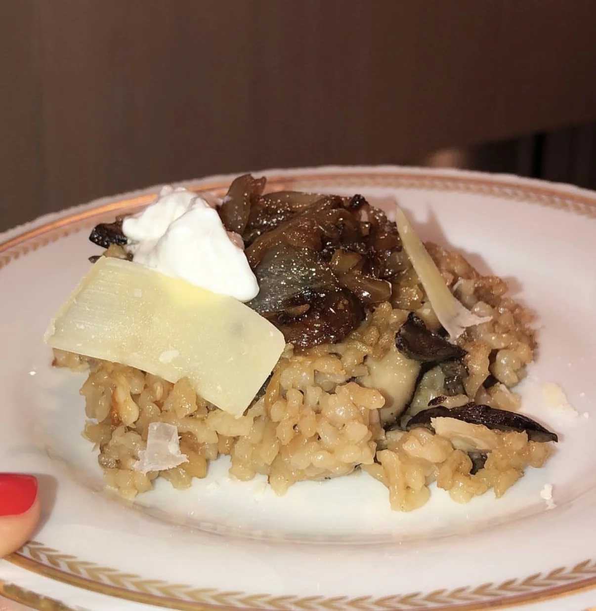 Close-up of a plate of mushroom risotto garnished with cheese, sour cream, and a red painted fingernail visible at the side.
