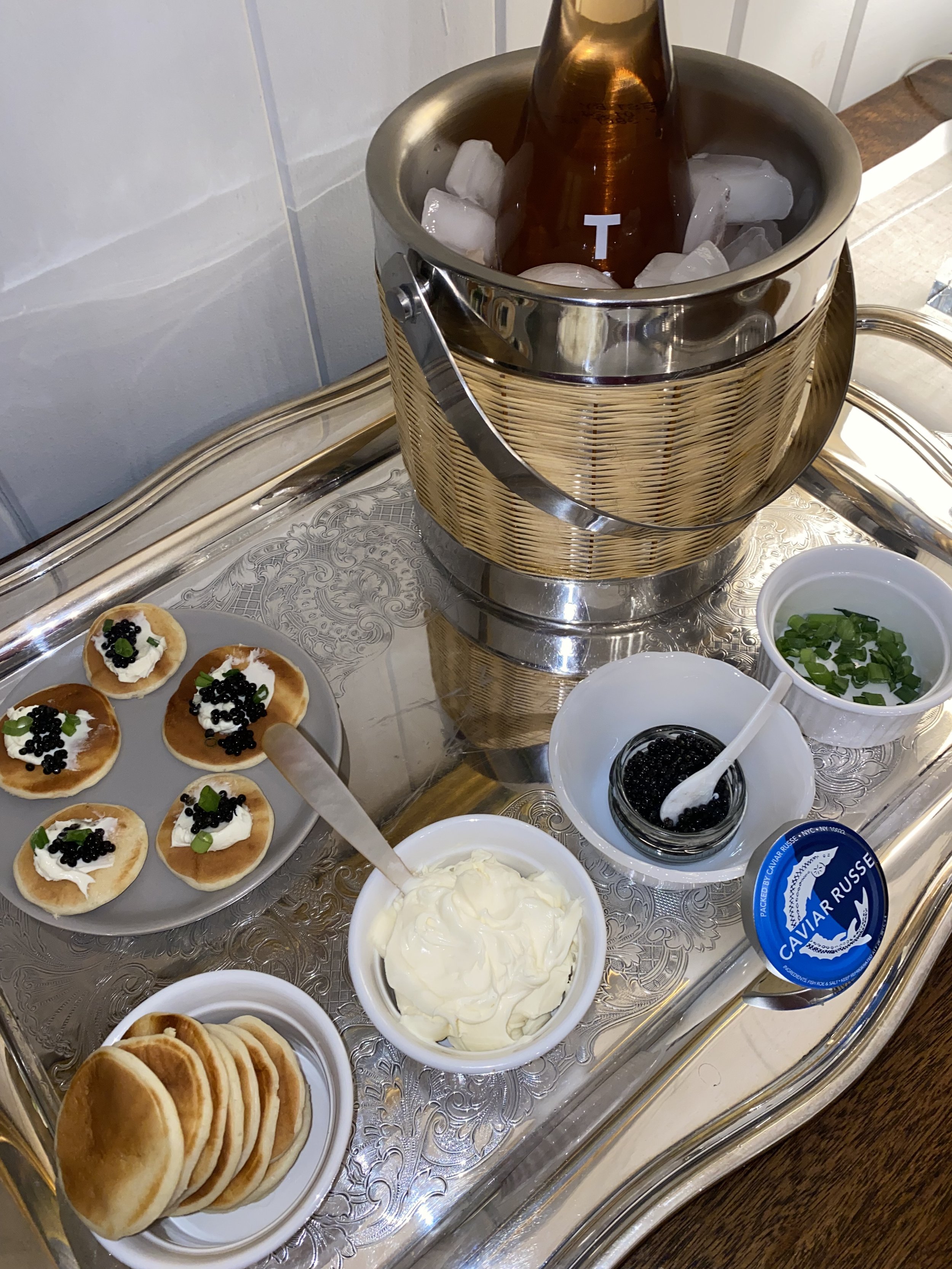 Tray with bowls of cream cheese, caviar, and garnishes, along with small pancakes topped with cream and caviar, and a bottle of champagne in an ice bucket.