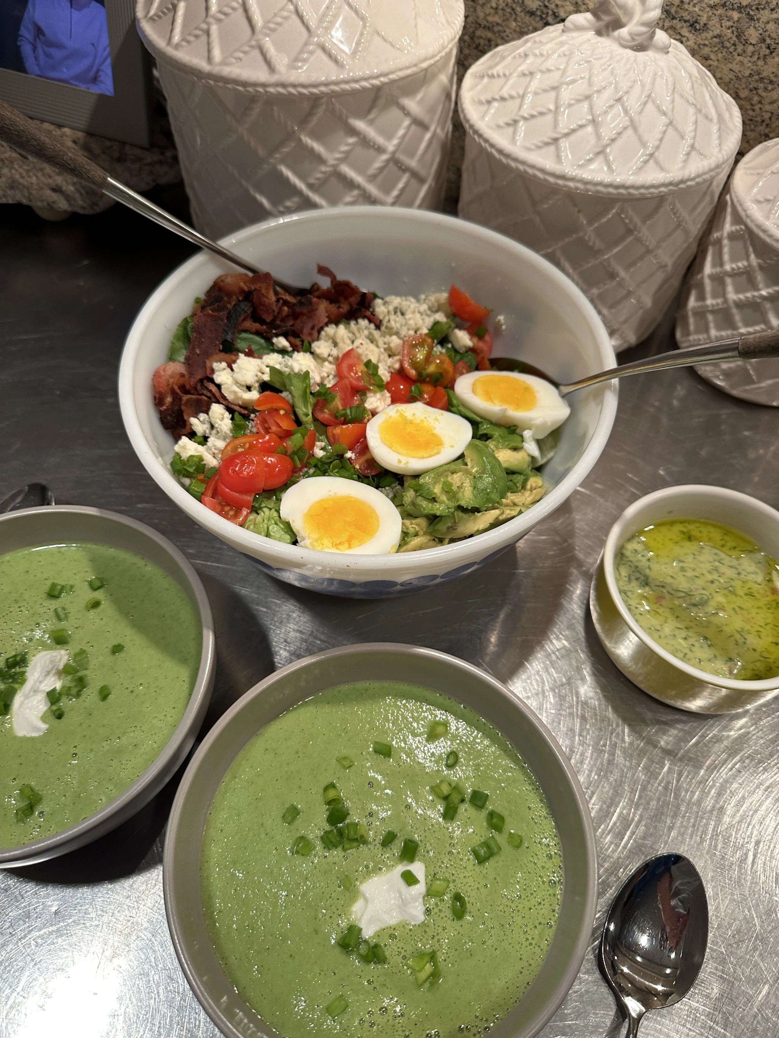 A bowl of salad with cherry tomatoes, chopped green onions, shredded cheese, bacon bits, and slices of hard-boiled eggs. Surrounding the salad are three bowls of green soup garnished with green onions and a dollop of sour cream, along with a small me