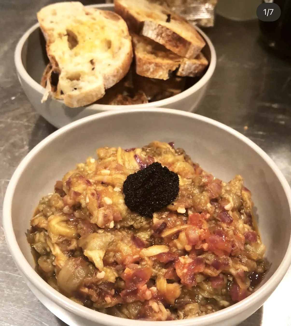 A bowl of chopped eggplant and tomato stew topped with black caviar, with a bowl of toasted bread slices behind it.