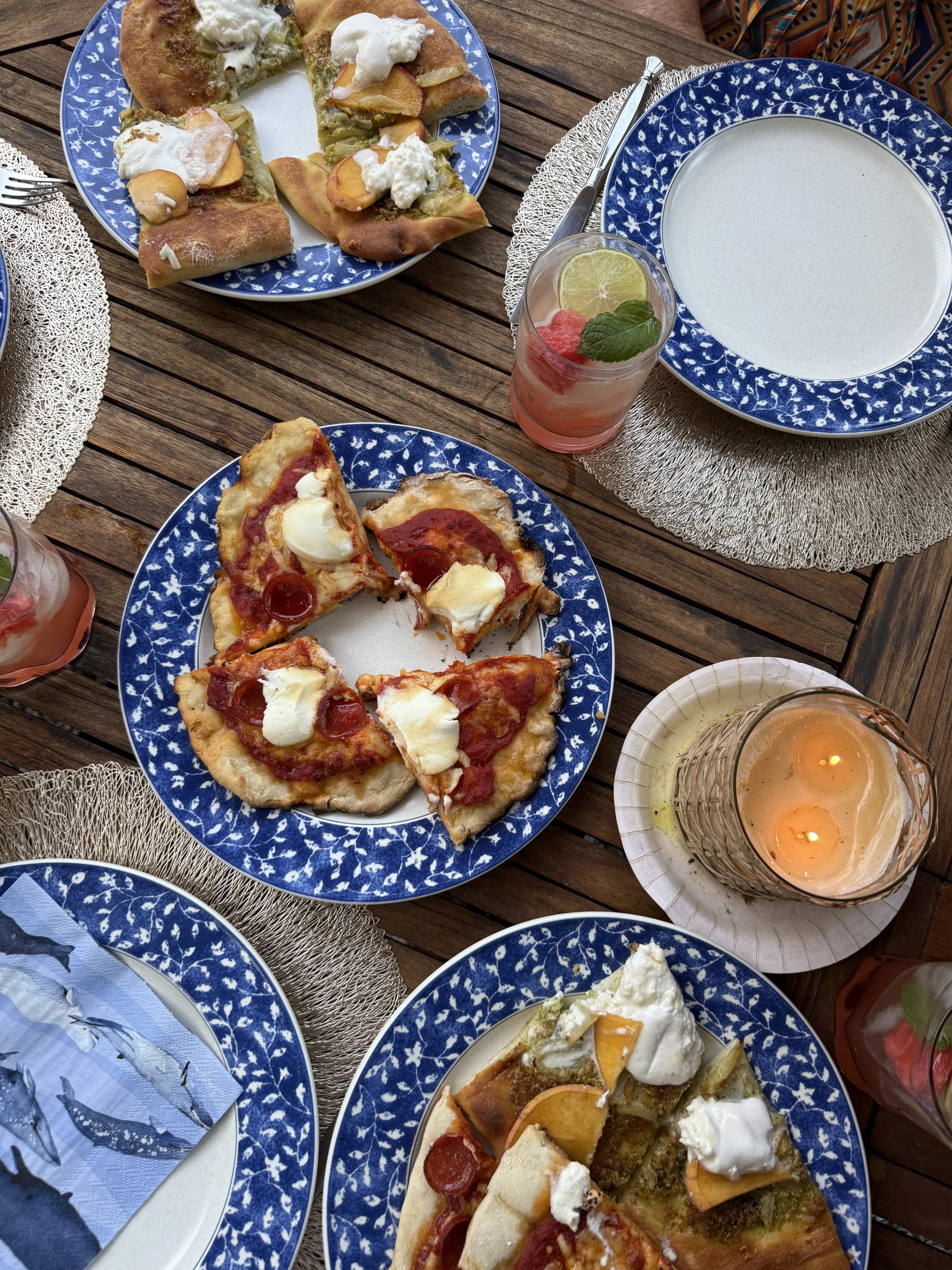 A table set with three plates of pizza, two glasses of pink drinks with strawberries and mint, and a candle holder with lit candles. The table has a wooden surface with woven placemats.