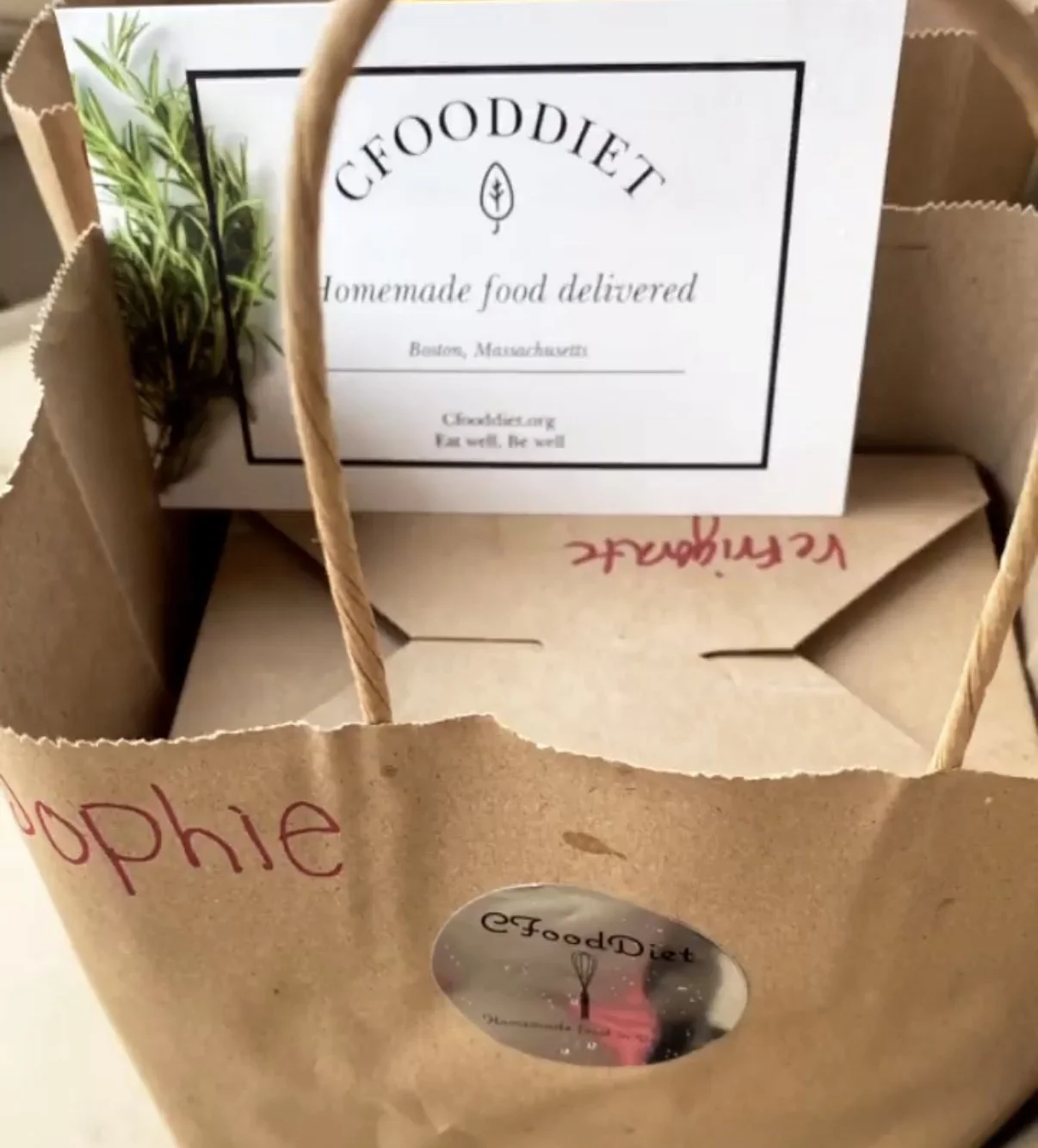 A brown paper bag with twisted paper handles, containing a sprig of rosemary and a card that says "GoodDiet: Homemade food delivered, Boston, Massachusetts, Cookbooking, Eat well, Be well."