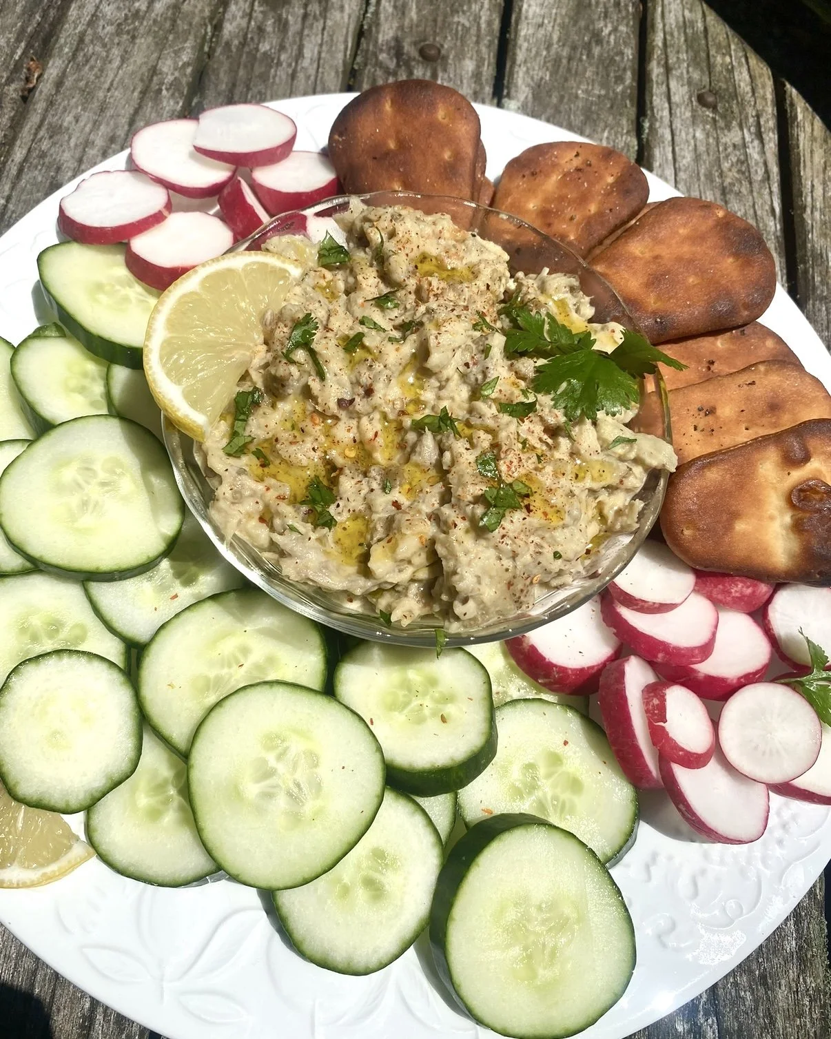 A plate of fresh sliced cucumbers, radishes, and lemon slices with a bowl of creamy eggplant dip garnished with parsley and olive oil, on a rustic wooden surface.