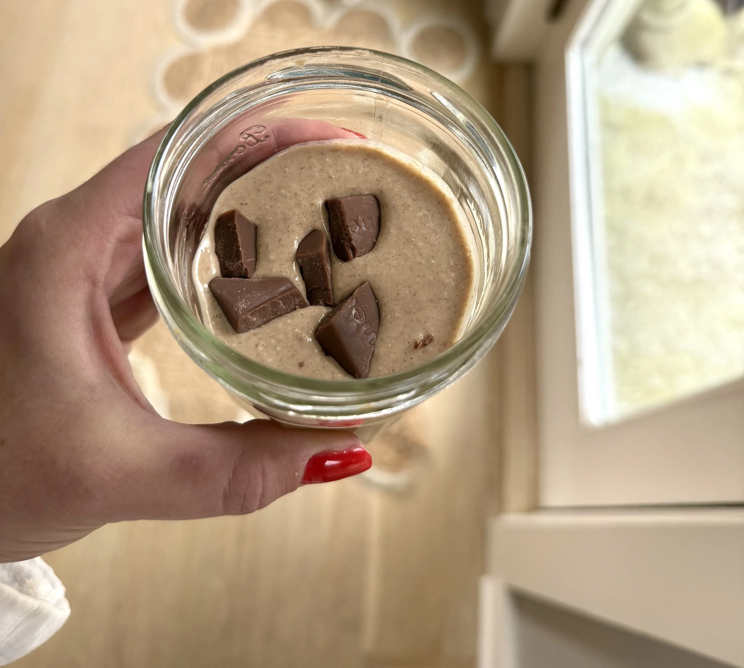 A hand with red nail polish holding a glass jar of creamy chocolate-hazelnut spread topped with broken chocolate pieces, near a window.