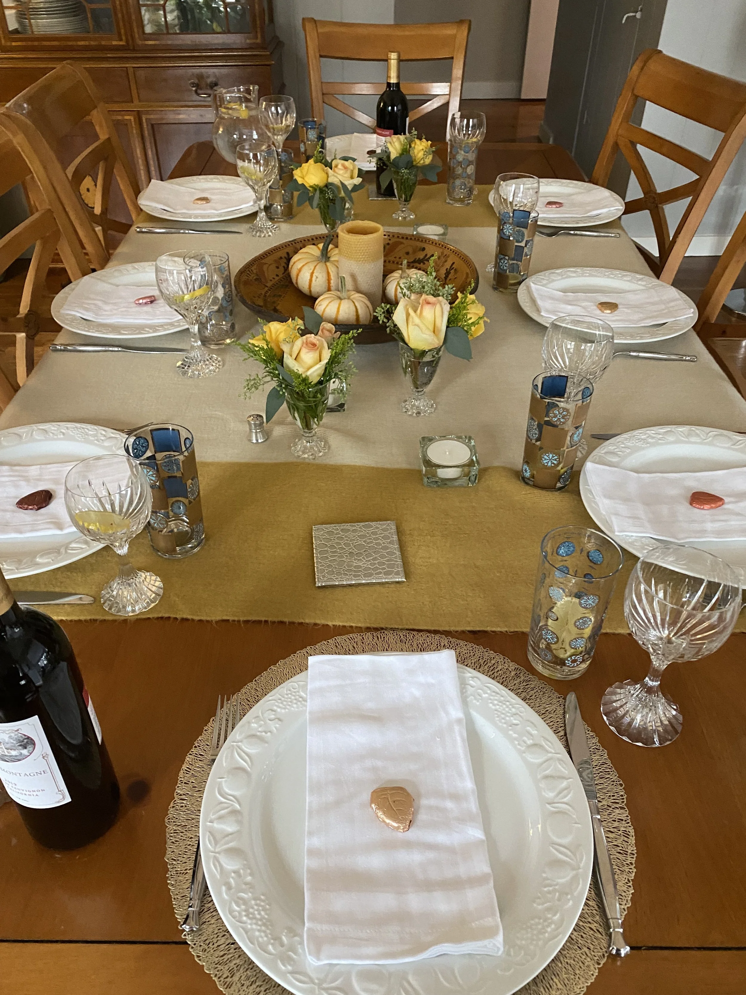 A dining table set for a meal with white plates, wine glasses, water glasses, utensils, and decorative centerpieces of yellow roses and small pumpkins. There is a bottle of wine in the center, and a gold-colored table runner with a small silver coile