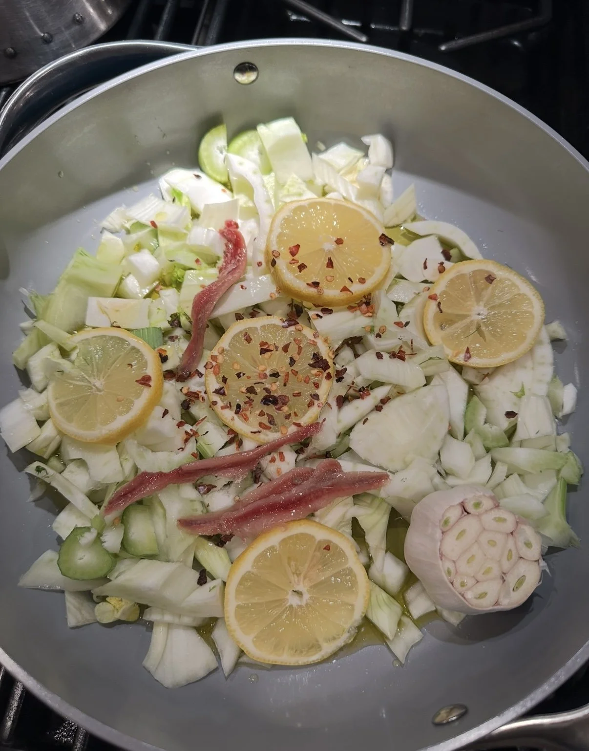 A stovetop pan with chopped cabbage, lemon slices sprinkled with chili flakes, garlic, and small pieces of fish or meat.