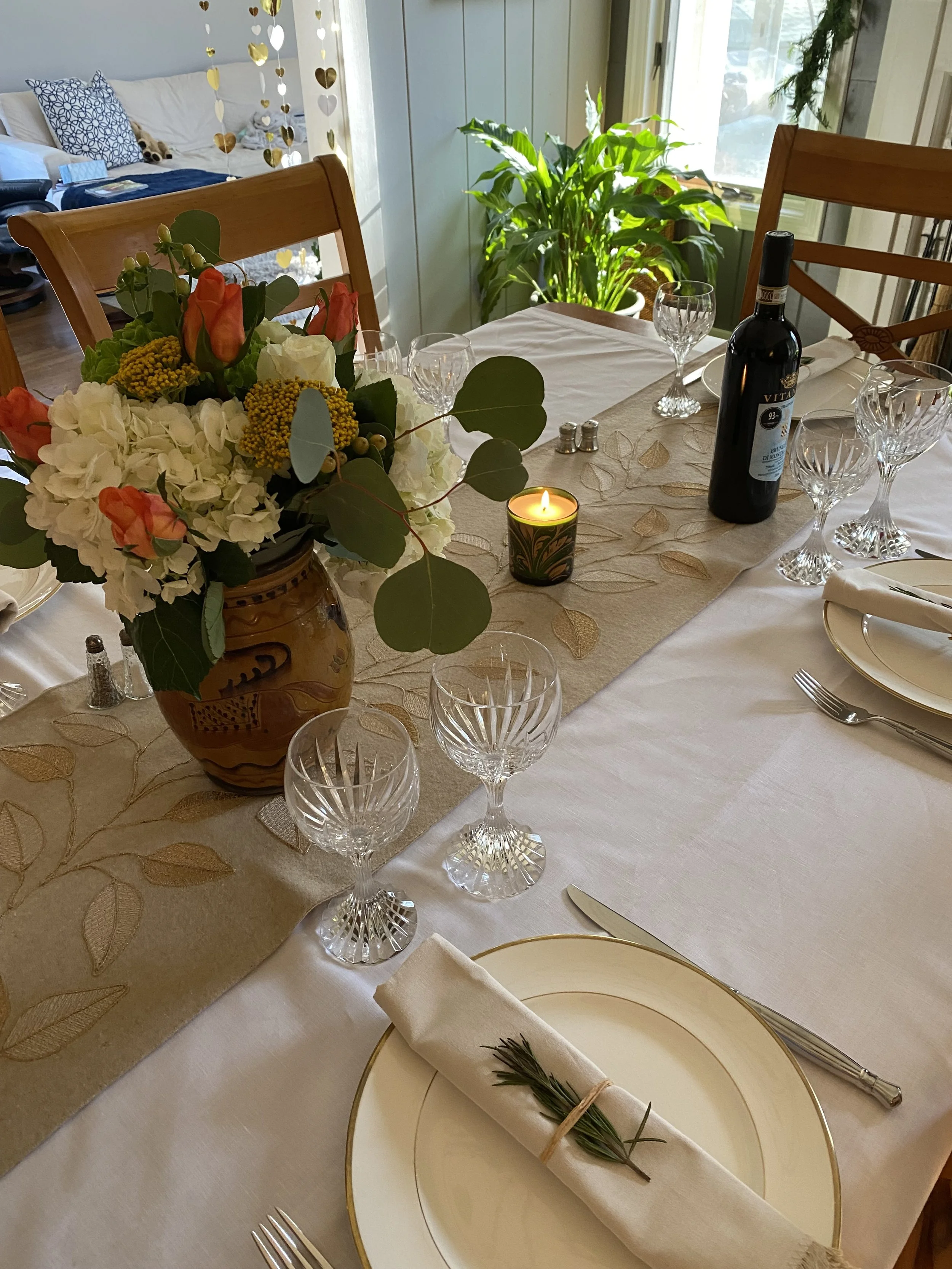 Elegant dining table set for a meal with a floral centerpiece, wine bottle, glassware, a candle, and place settings including a napkin with a sprig of rosemary.