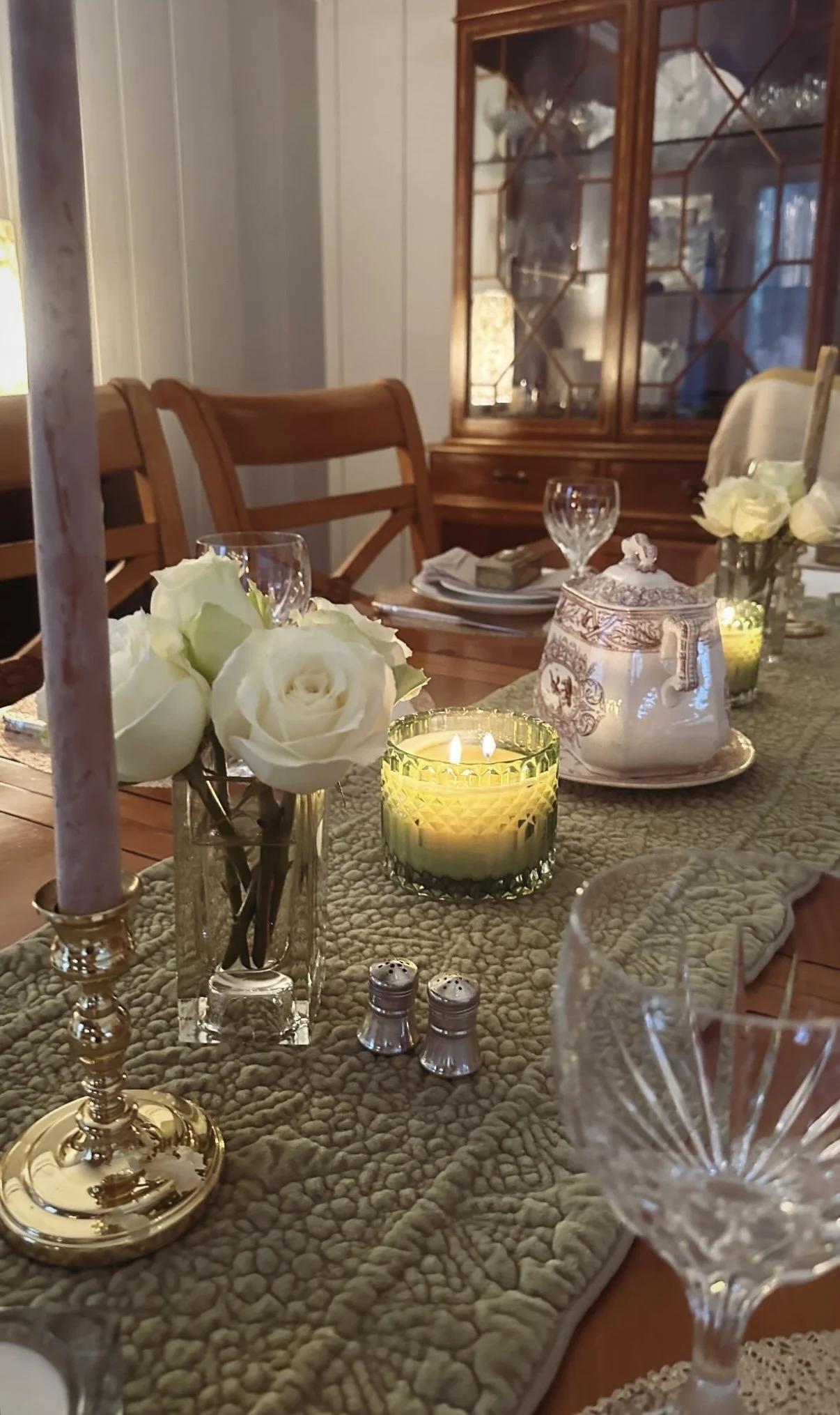 A dining table decorated with white roses in a glass vase, lit candles, a teapot with floral patterns, salt and pepper shakers, and crystal glassware in a cozy, warmly lit room.