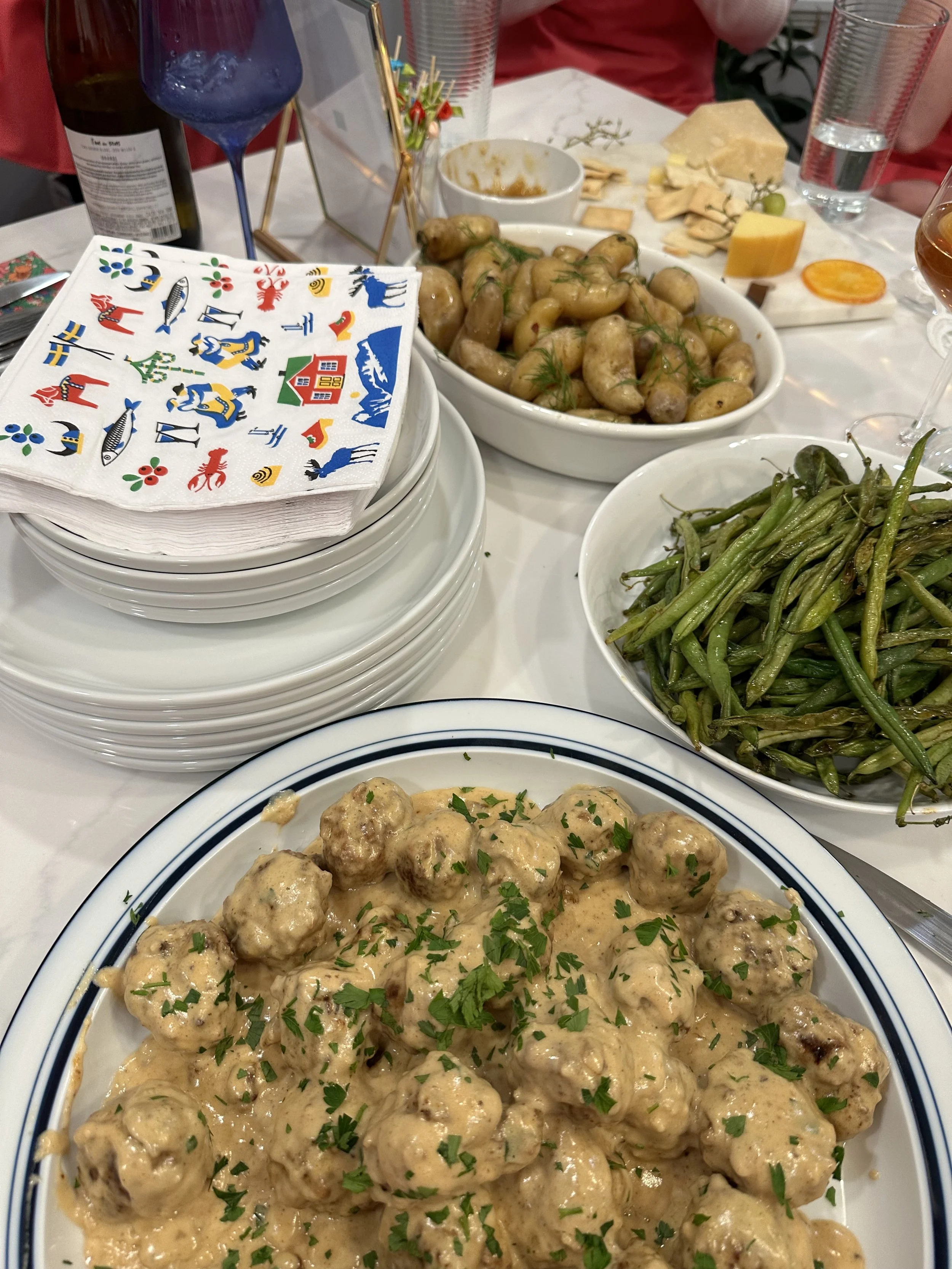A table full of various Swedish dishes including meatballs with creamy gravy, green beans, boiled potatoes, and lingonberry sauce. There are multiple glasses and a large stack of white plates.