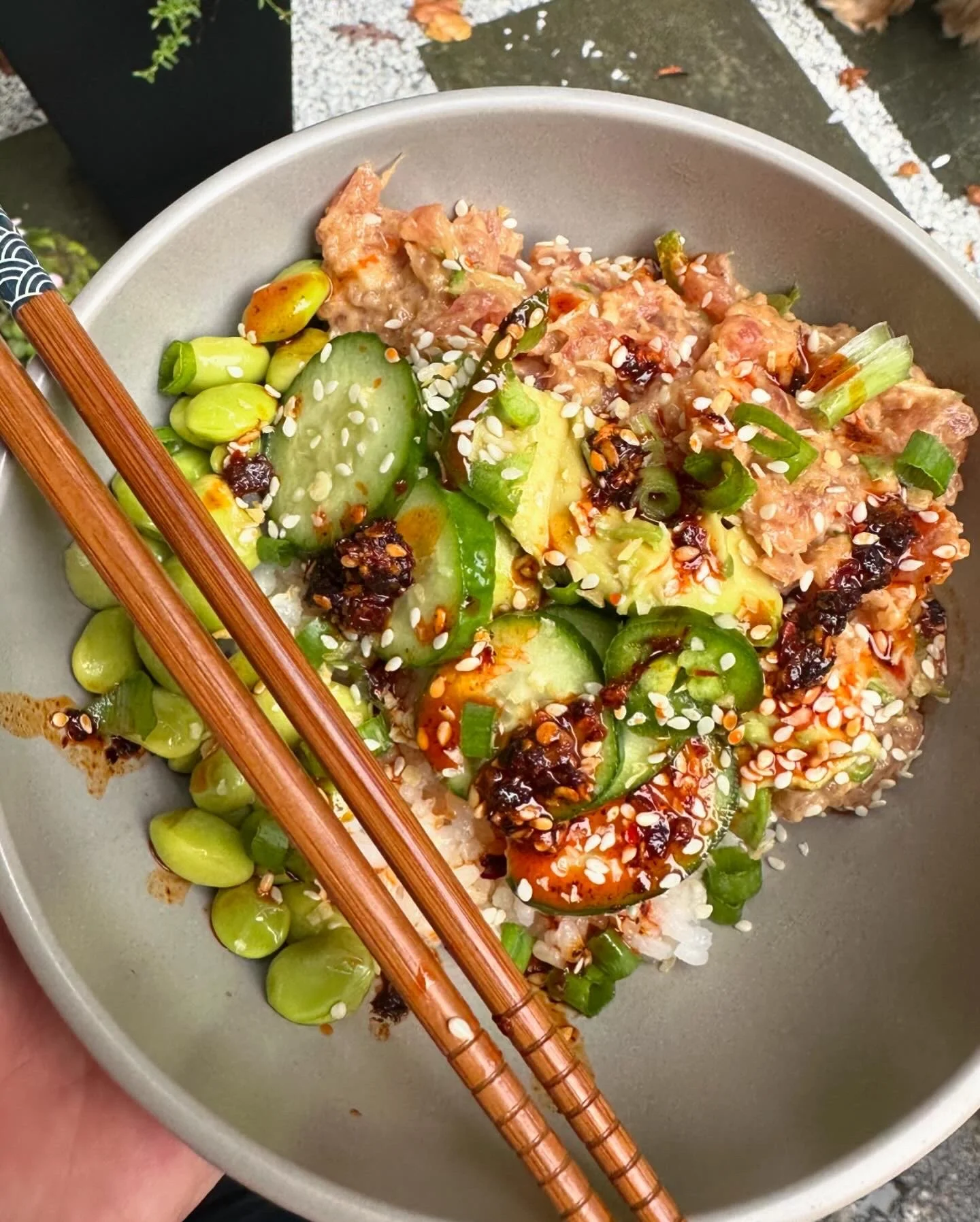 A bowl of rice topped with sliced cucumbers, chopped green onions, sesame seeds, and drizzled with dark soy sauce and chili oil, with chopsticks resting on the side.