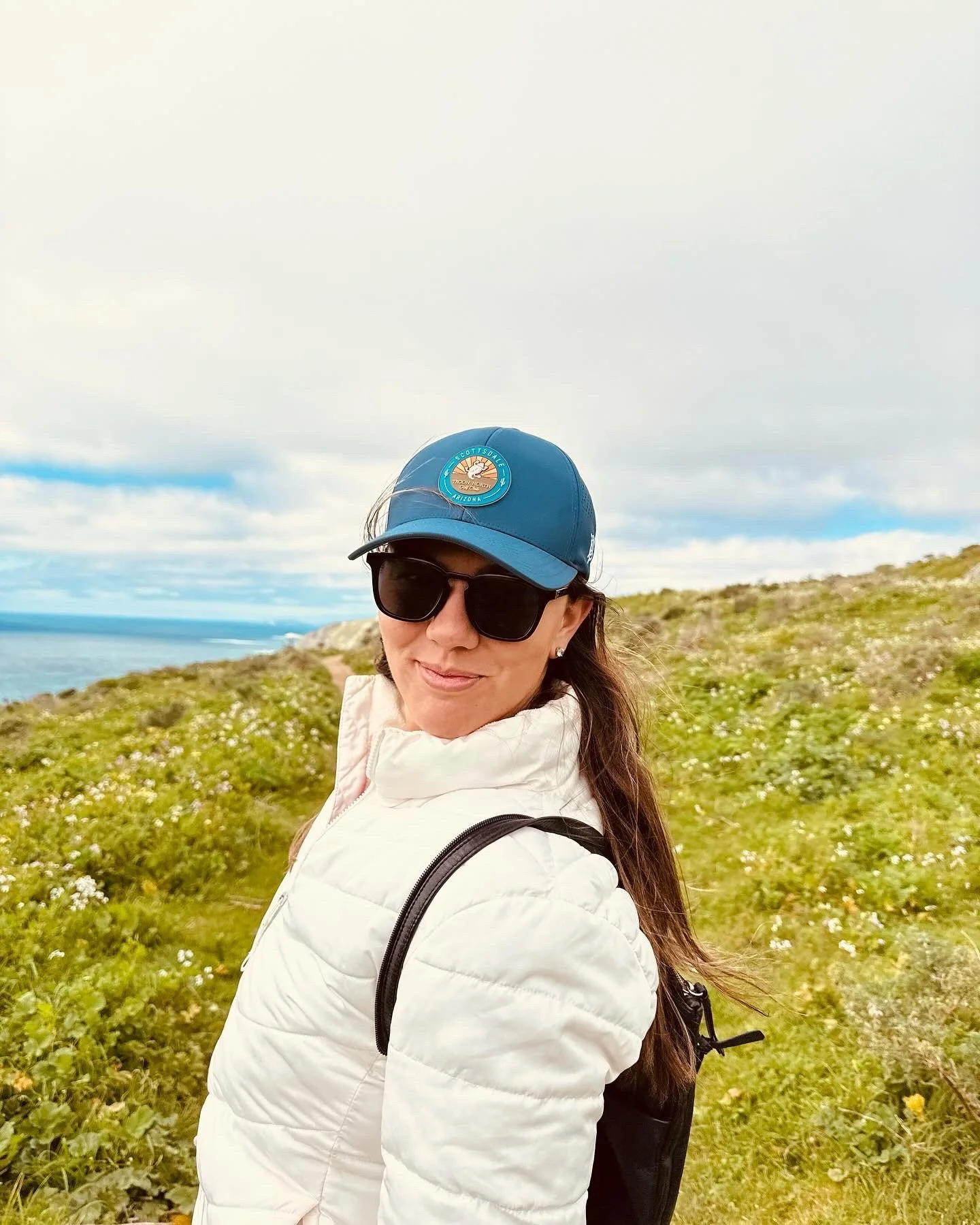 Woman wearing sunglasses, a blue cap, and a white jacket standing outdoors on a grassy hill with flowers, overlooking the ocean and cloudy sky.