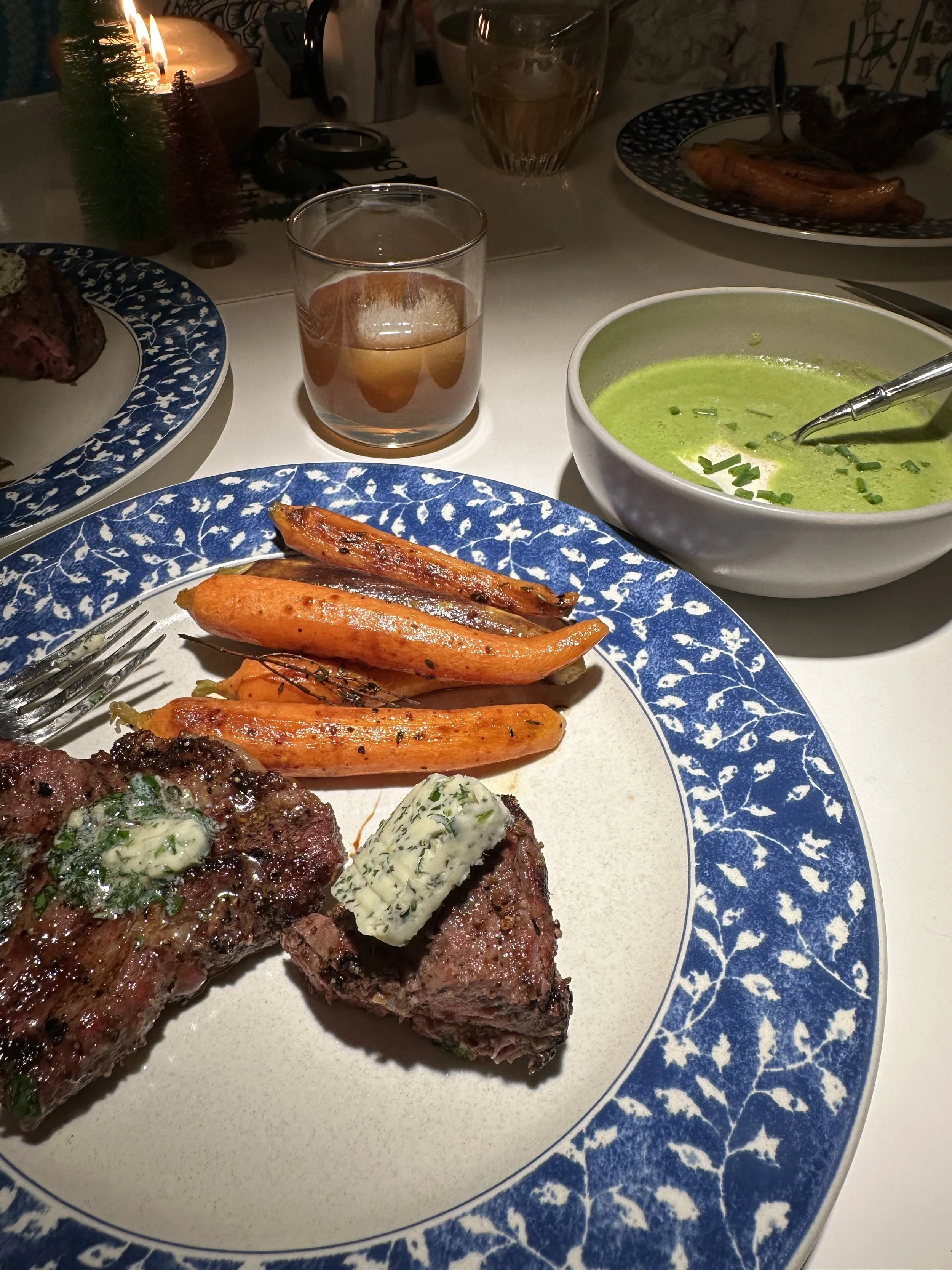 Dinner plate with cooked steak topped with herb mixture and butter, roasted carrots, and sweet potato fries. Additional bowl of green soup, a glass of white wine, and a glass of rosé wine on the table.