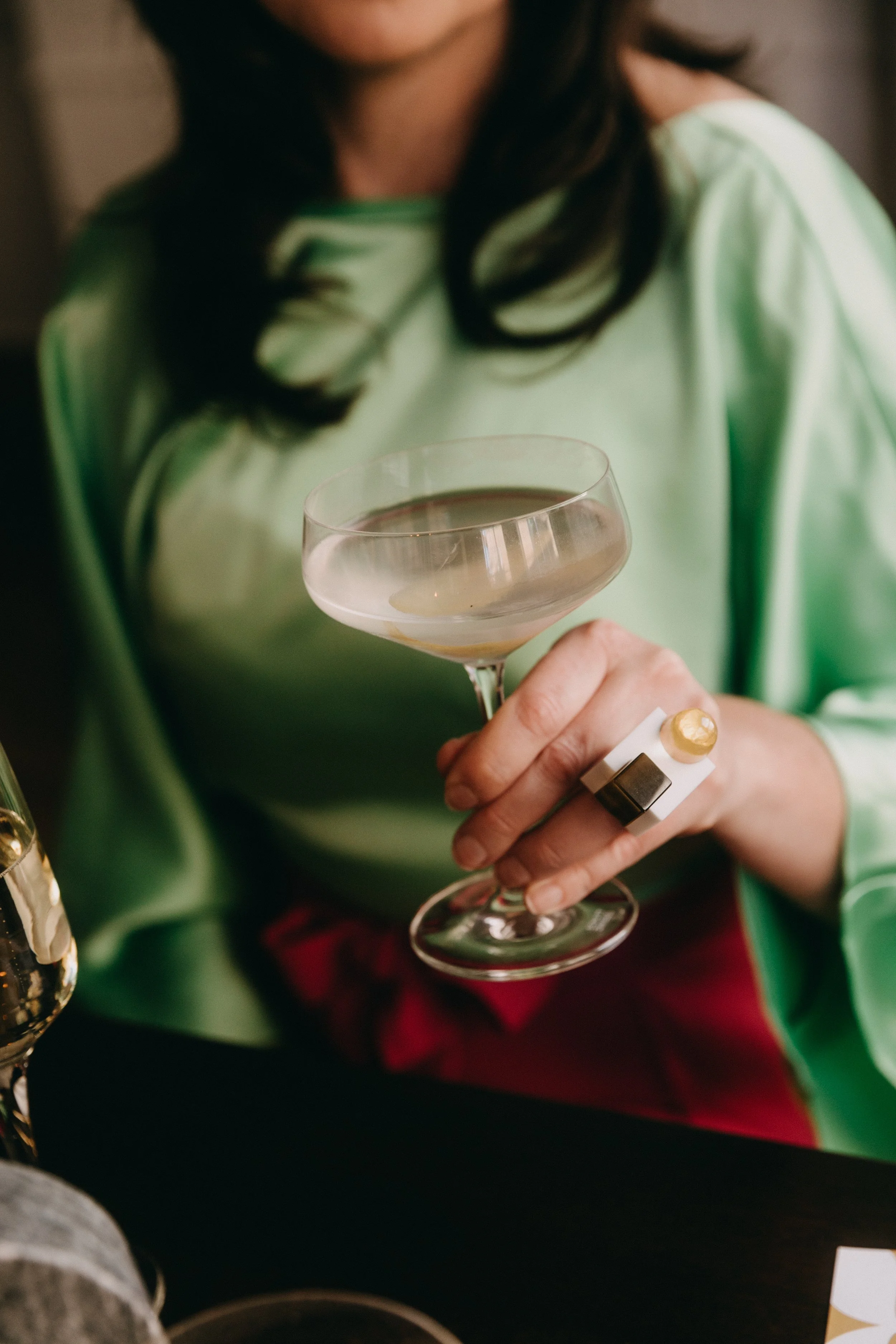 Person holding a cocktail glass with a clear drink, wearing a ring with gold and black accents, dressed in a green and white top, during a small wedding or celebration.