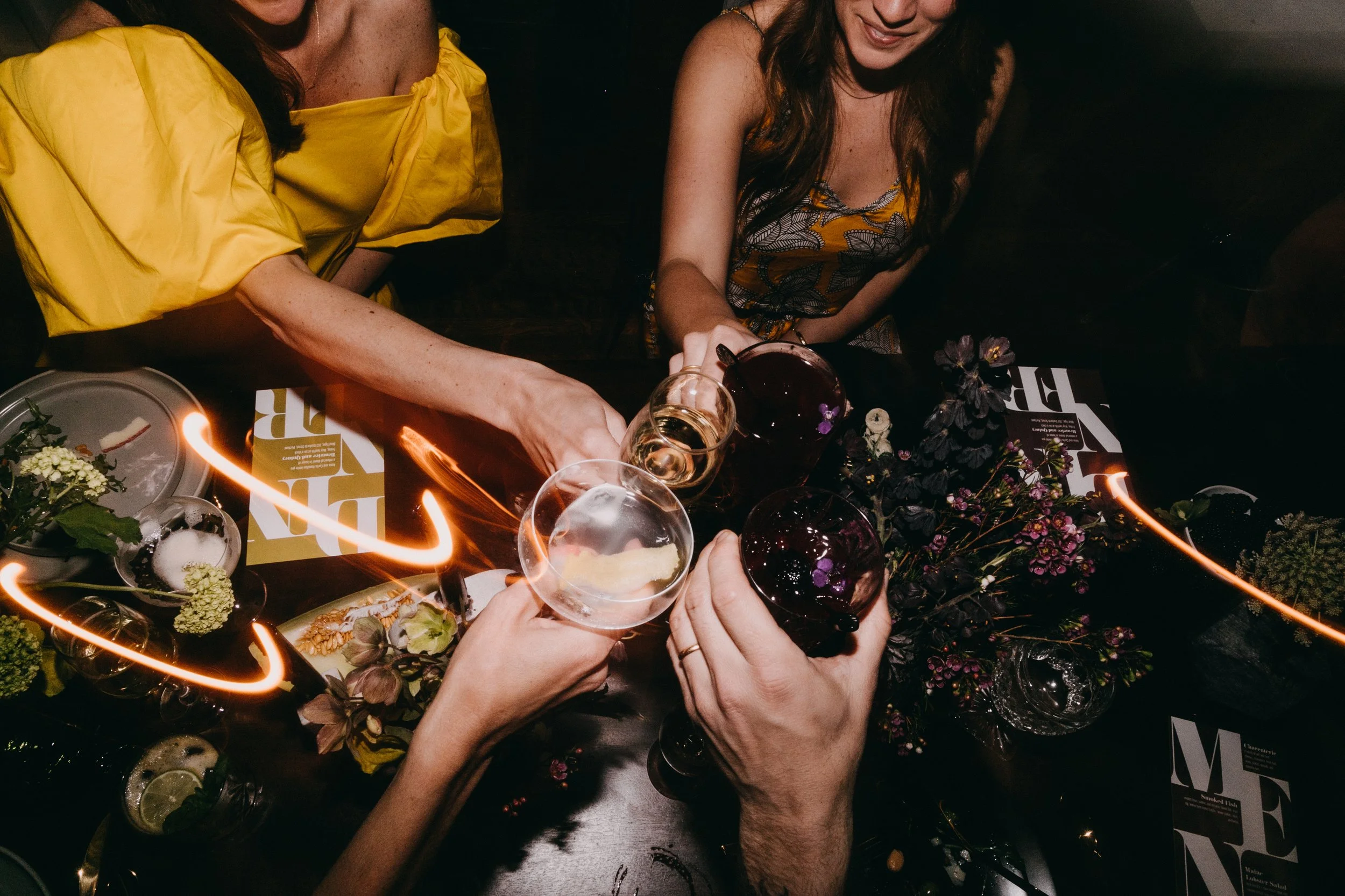 People toasting with glasses of wine and cocktails at a table decorated with flowers and candles, during a wedding or elopement.