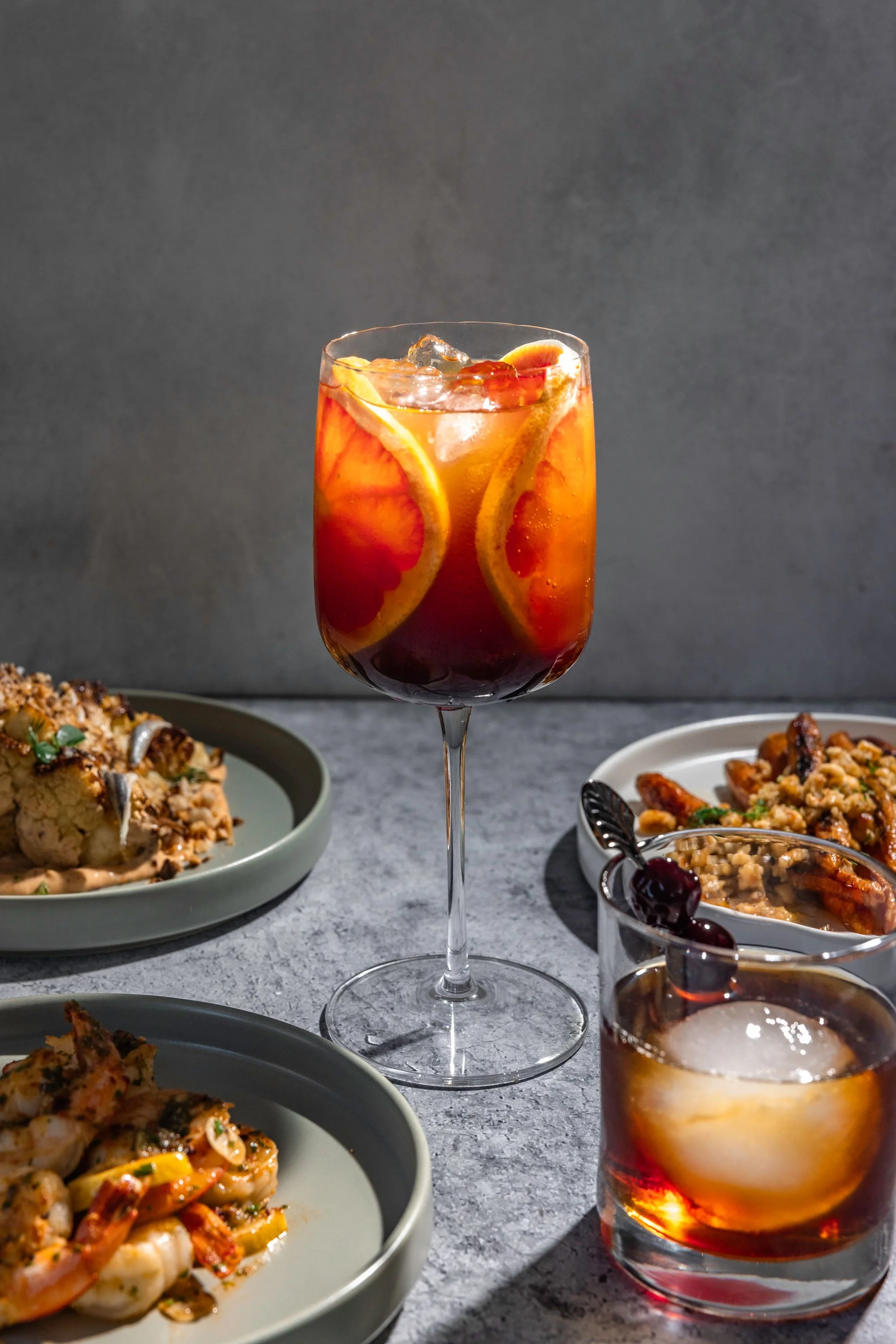 A red cocktail in a wine glass with orange slices, along with plates of food including shrimp and rice, on a gray textured surface.