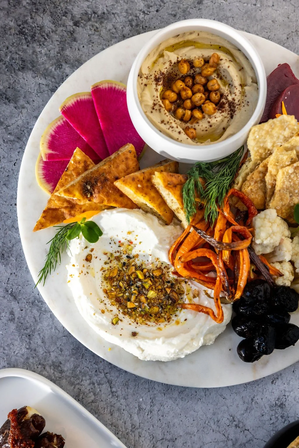 A plate with Mediterranean appetizers, including a bowl of hummus topped with chickpeas, radish slices, toasted pita chips, a scoop of yogurt garnished with herbs and seeds, roasted vegetables, cauliflower, and black olives.