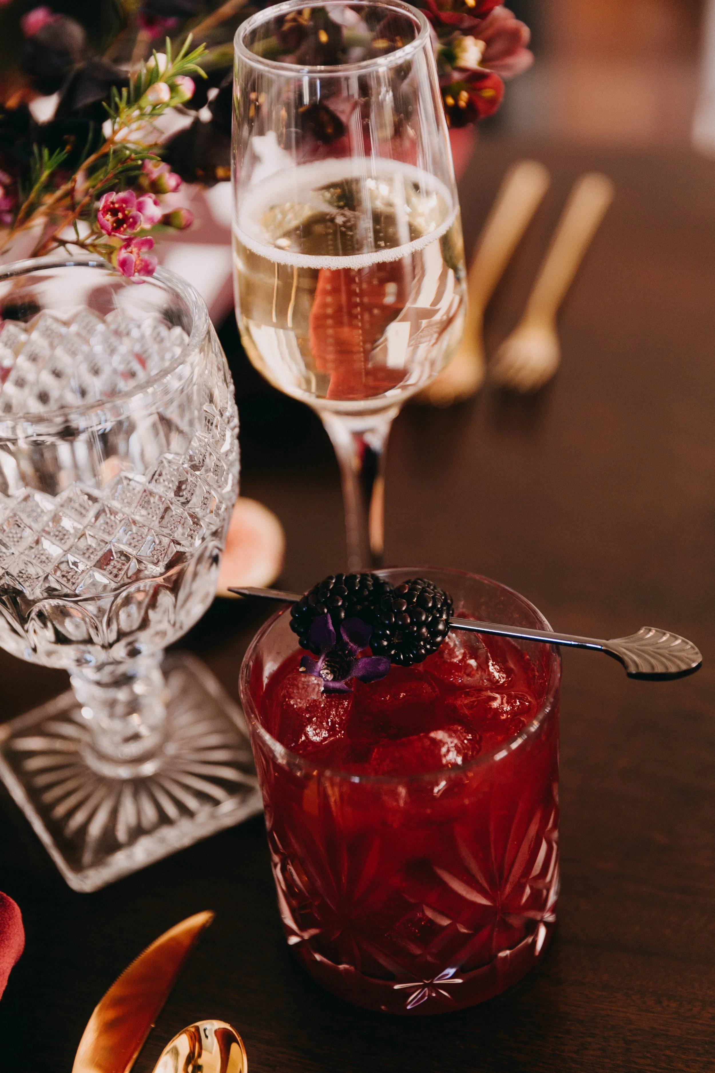 A glass of champagne and a red cocktail with blackberries on top on a dark table, surrounded by crystal glassware, flowers, and gold utensils.