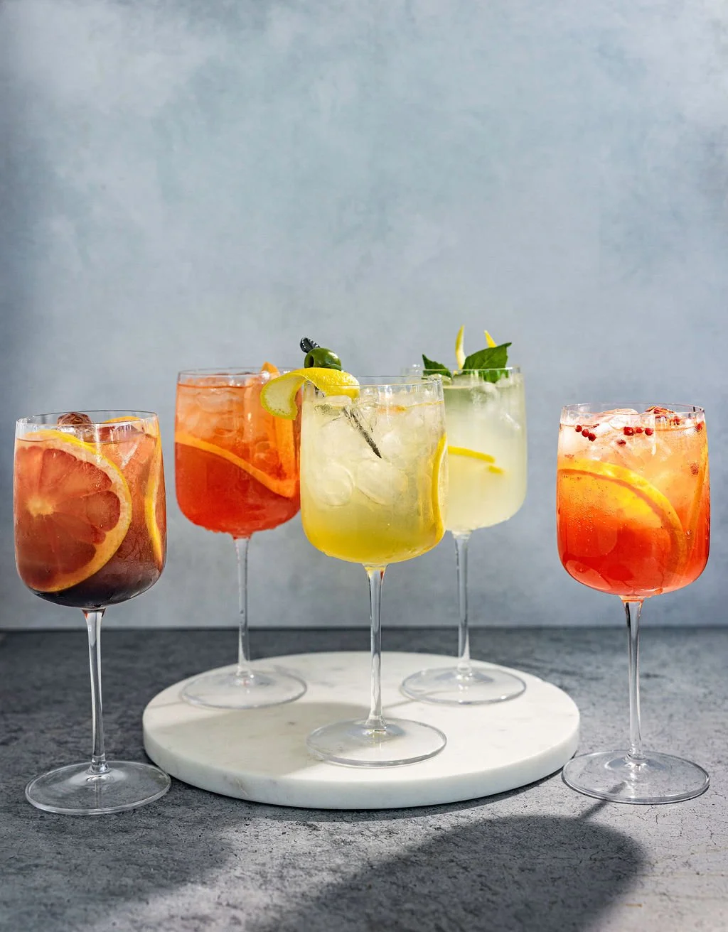Five colorful craft cocktails in wine glasses on a white marble tray against a neutral background.