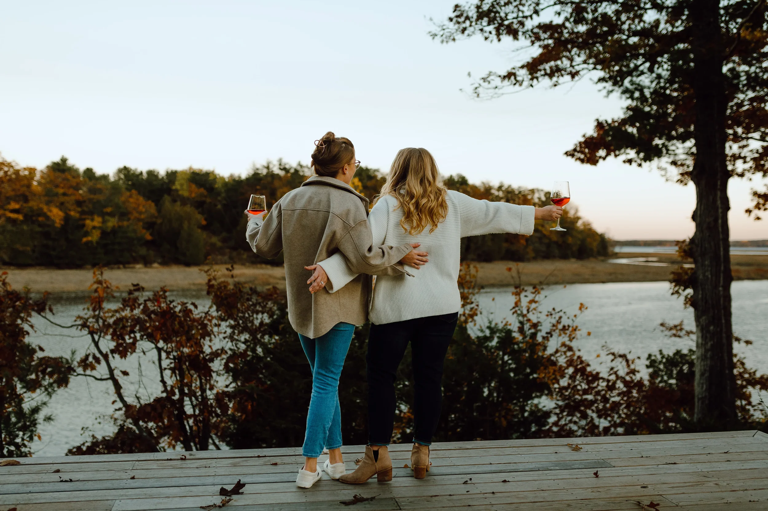 Two women stand on a dock near a lake at sunset, holding glasses of rosé wine, with autumn trees in the background.
