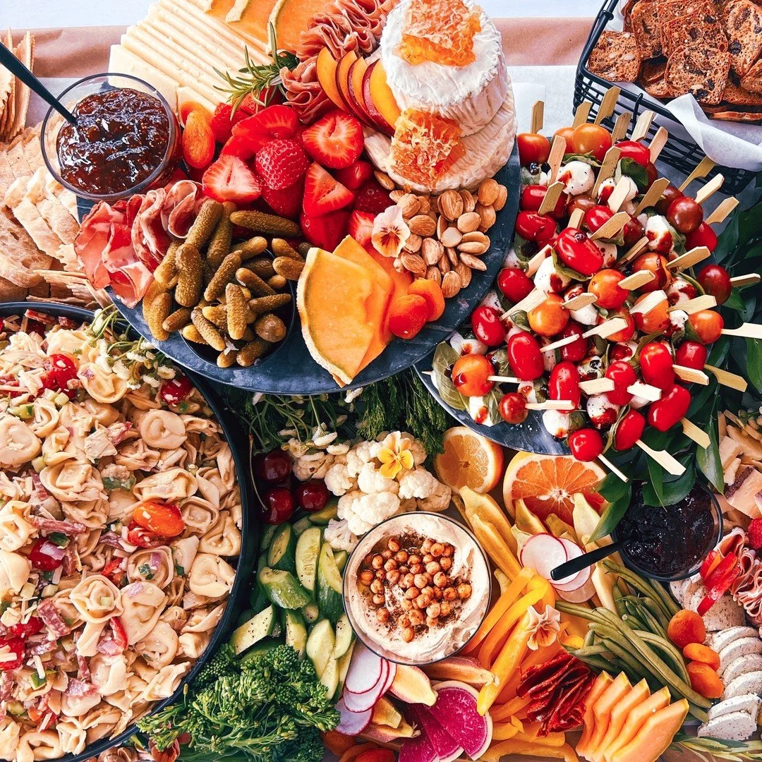 A colorful spread of foods including strawberries, sliced melons, grapes, cherry tomatoes, roasted nuts, pepperoni, bread, pasta salad, pickles, cauliflower, radishes, carrot sticks, and a bowl of hummus, with drinks, in a festive setting.