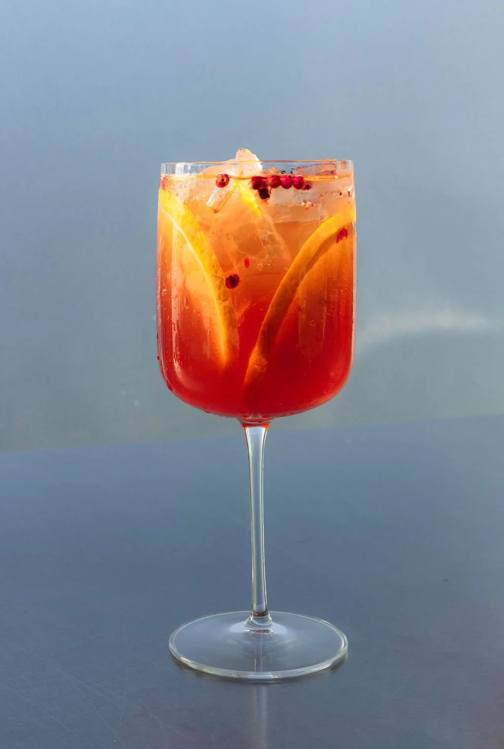 A red craft cocktail with orange slices and red berries, served over ice in a wine glass on a gray surface with a blue-gray background.