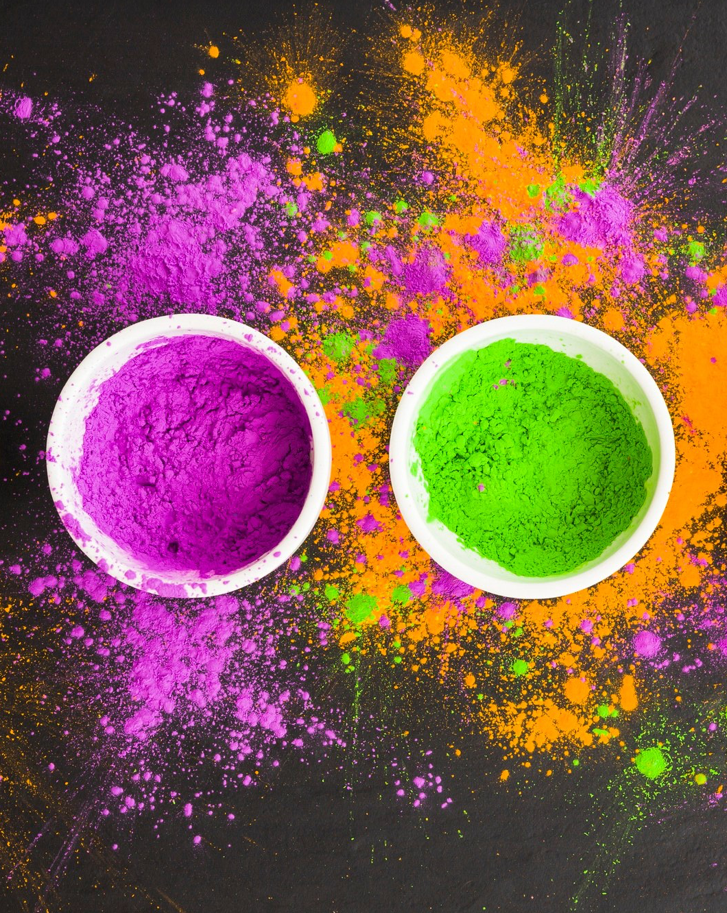 Zwei Schalen mit pinker und grüner Pulverfarbe auf einer schwarzen Oberfläche, umgeben von verstreuten farbigen Spritzern und Staubpartikeln