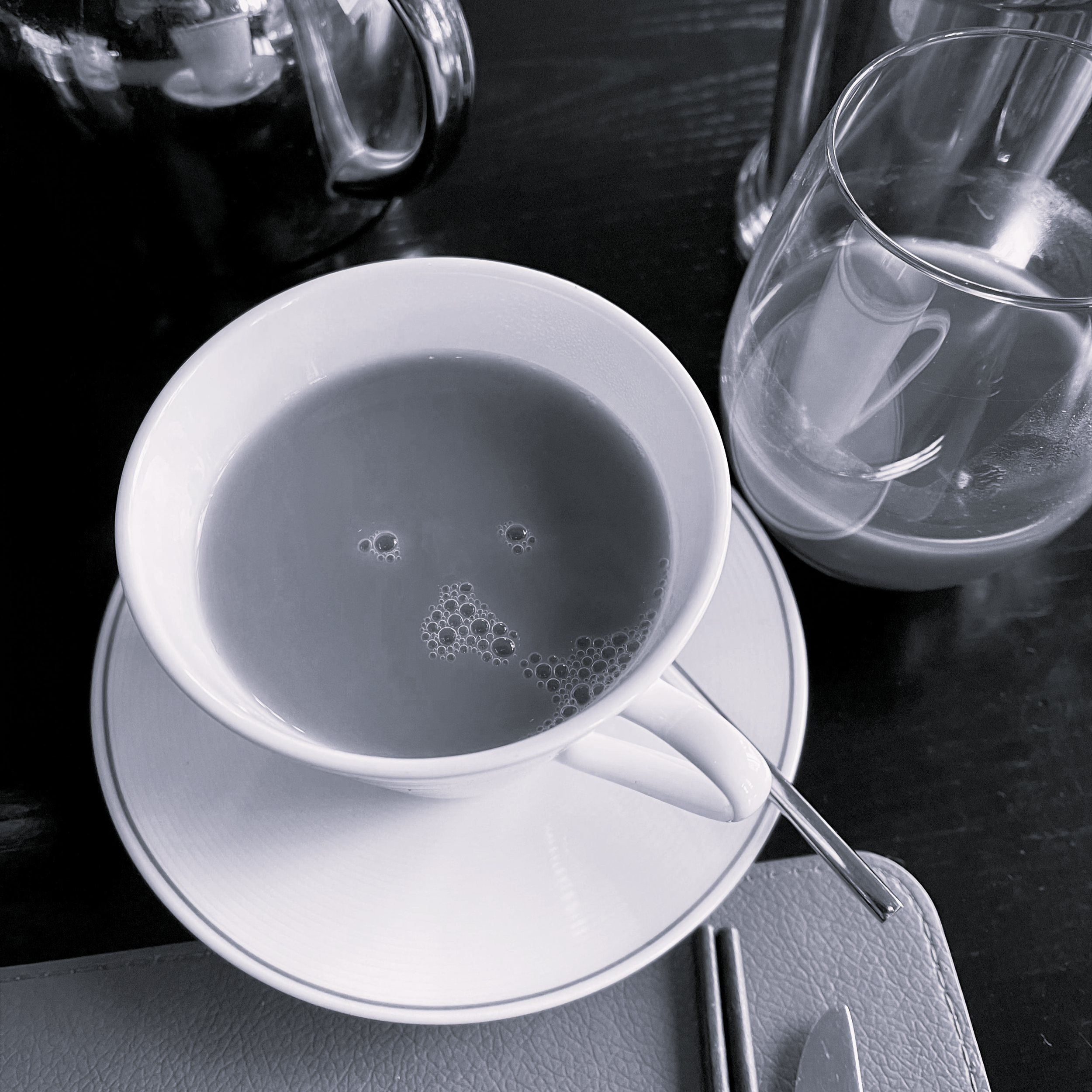 A face of bubbles in tea within a white coffee cup filled with a dark beverage, placed on a matching saucer, with a spoon resting on the saucer's edge. To the right, there is a transparent glass with a beverage.