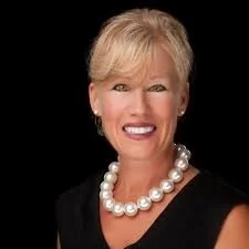 A smiling woman with short blonde hair wearing a black top and a pearl necklace.