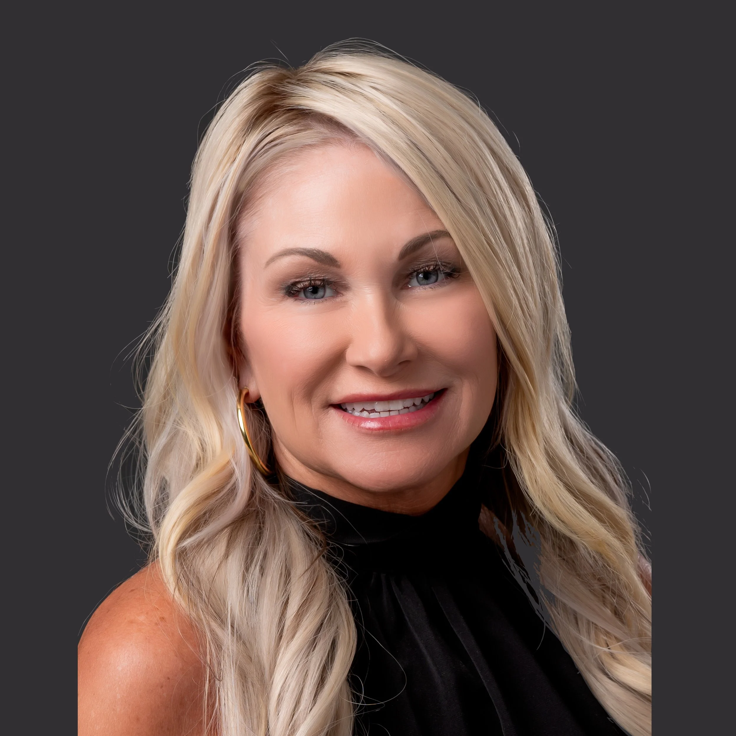 A portrait of a smiling blonde woman with blue eyes, wearing hoop earrings and a black top against a dark background.