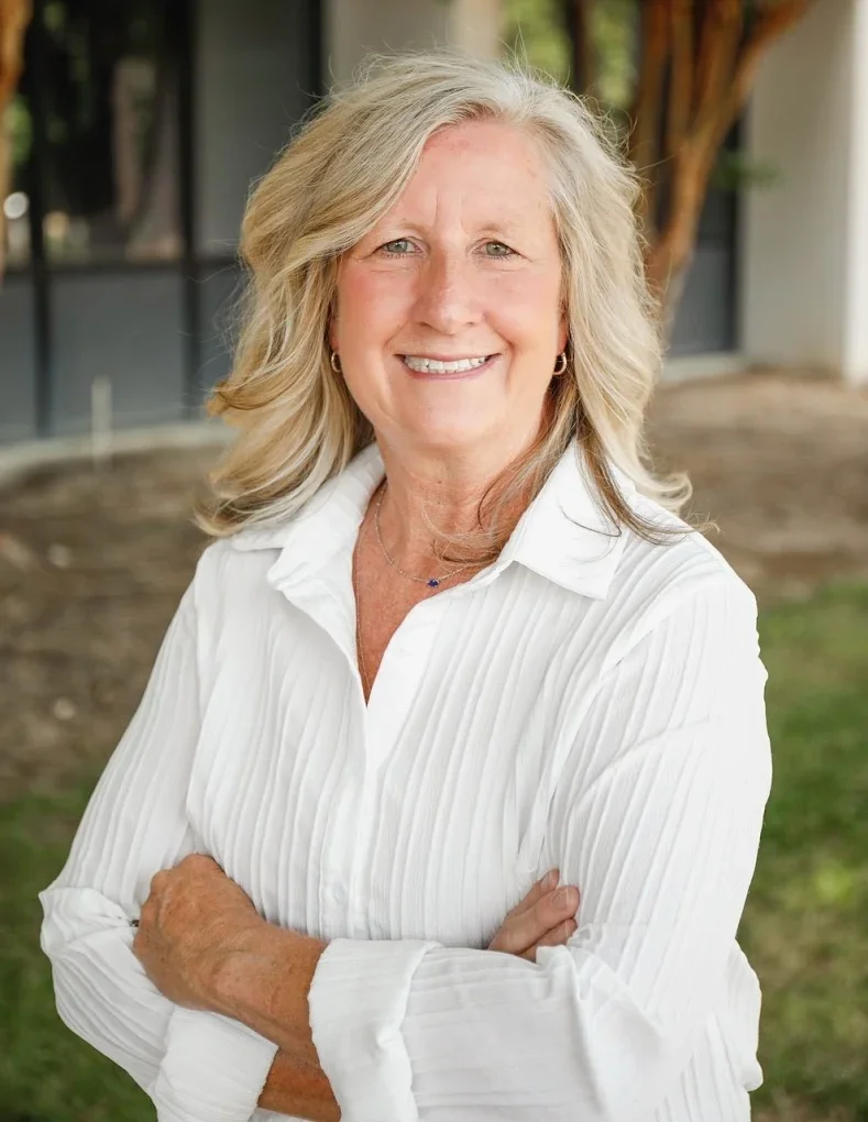 A smiling middle-aged woman with blonde hair wearing a white long-sleeve blouse, standing outdoors with a blurred background of a building, trees, and grass.