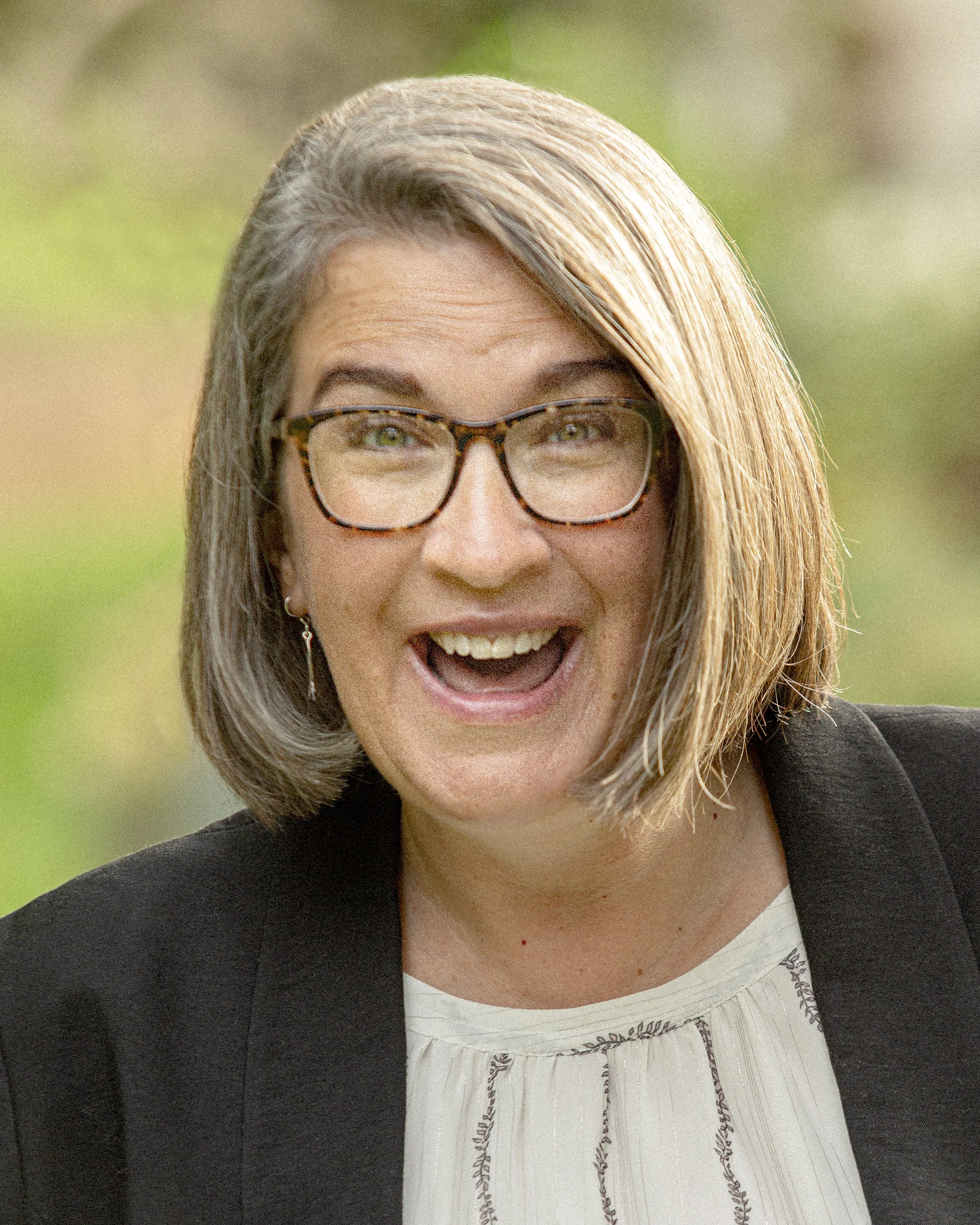 A woman with shoulder-length blonde hair, glasses, and earrings, smiling outdoors with a blurred green background.
