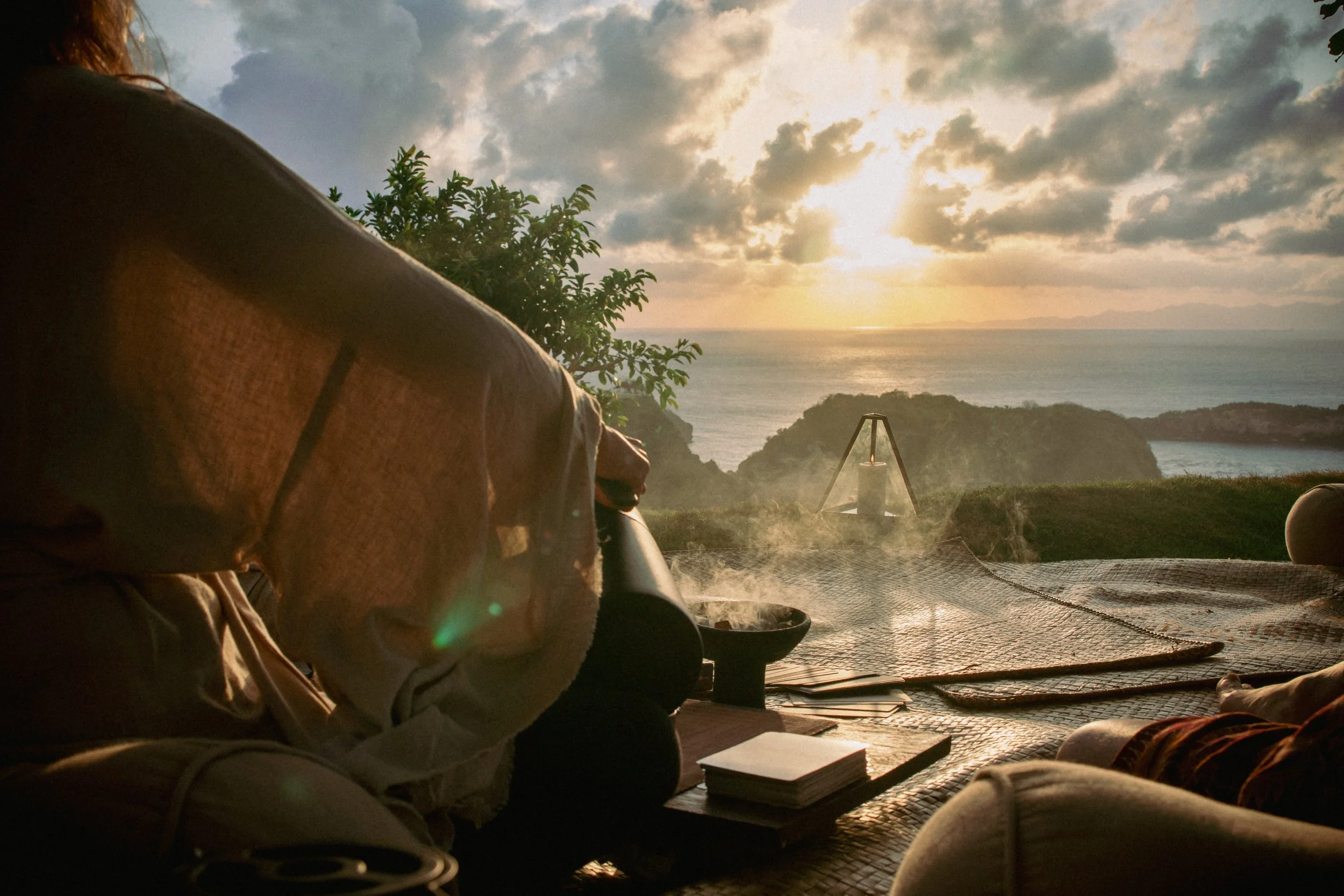 People relaxing outdoors at sunset, overlooking the ocean with clouds and mountains in the distance, sitting on a blanket with books and a lantern nearby.