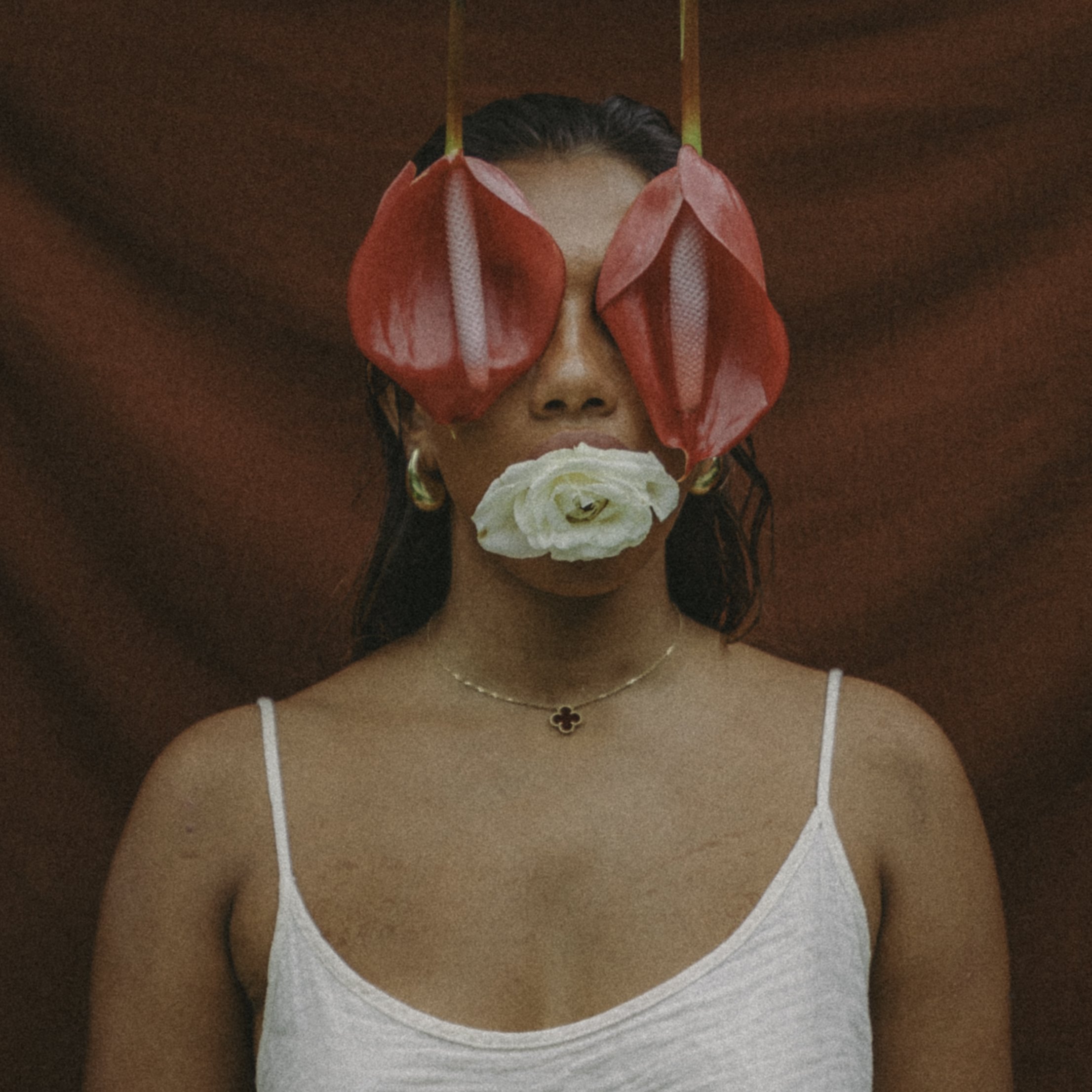 Woman with floral face covering, wearing large red leaf-like glasses and a white tank top, against a brown background.