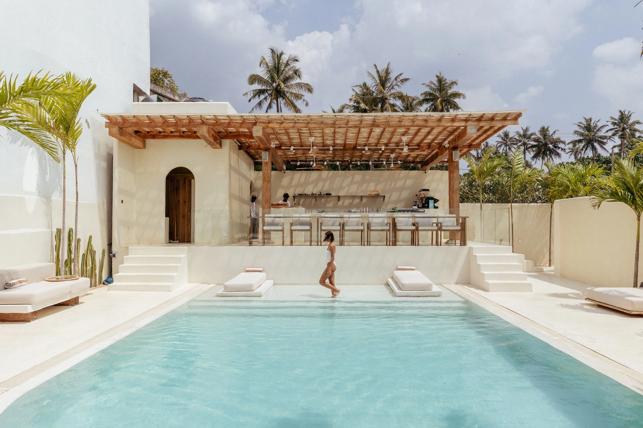 A woman walking in a swimming pool at a modern outdoor bar area with white furniture, palm trees, and an open sky.