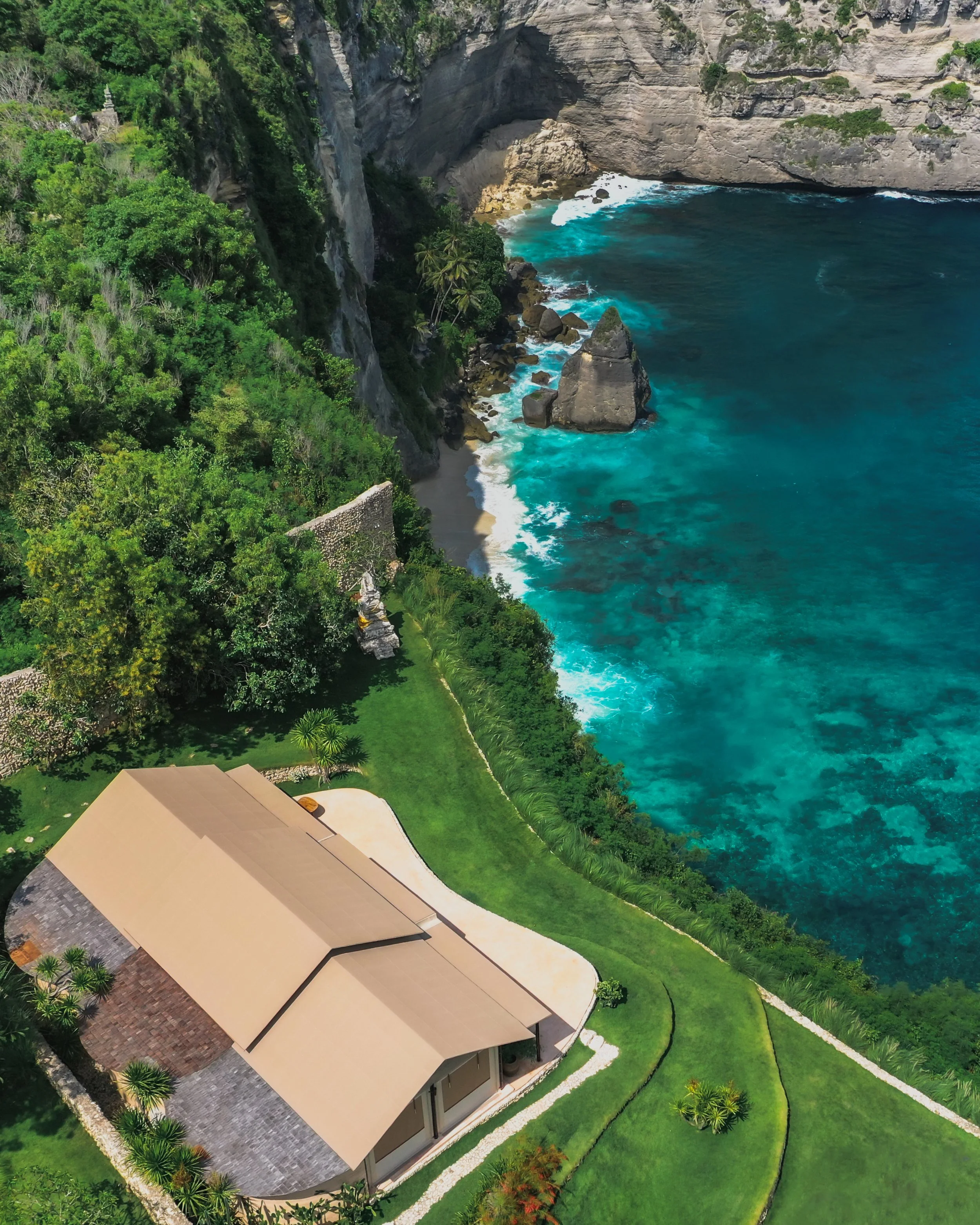 A house with a curved beige roof surrounded by green grass, with a cliffside and lush trees overlooking a turquoise ocean with rocks and waves.